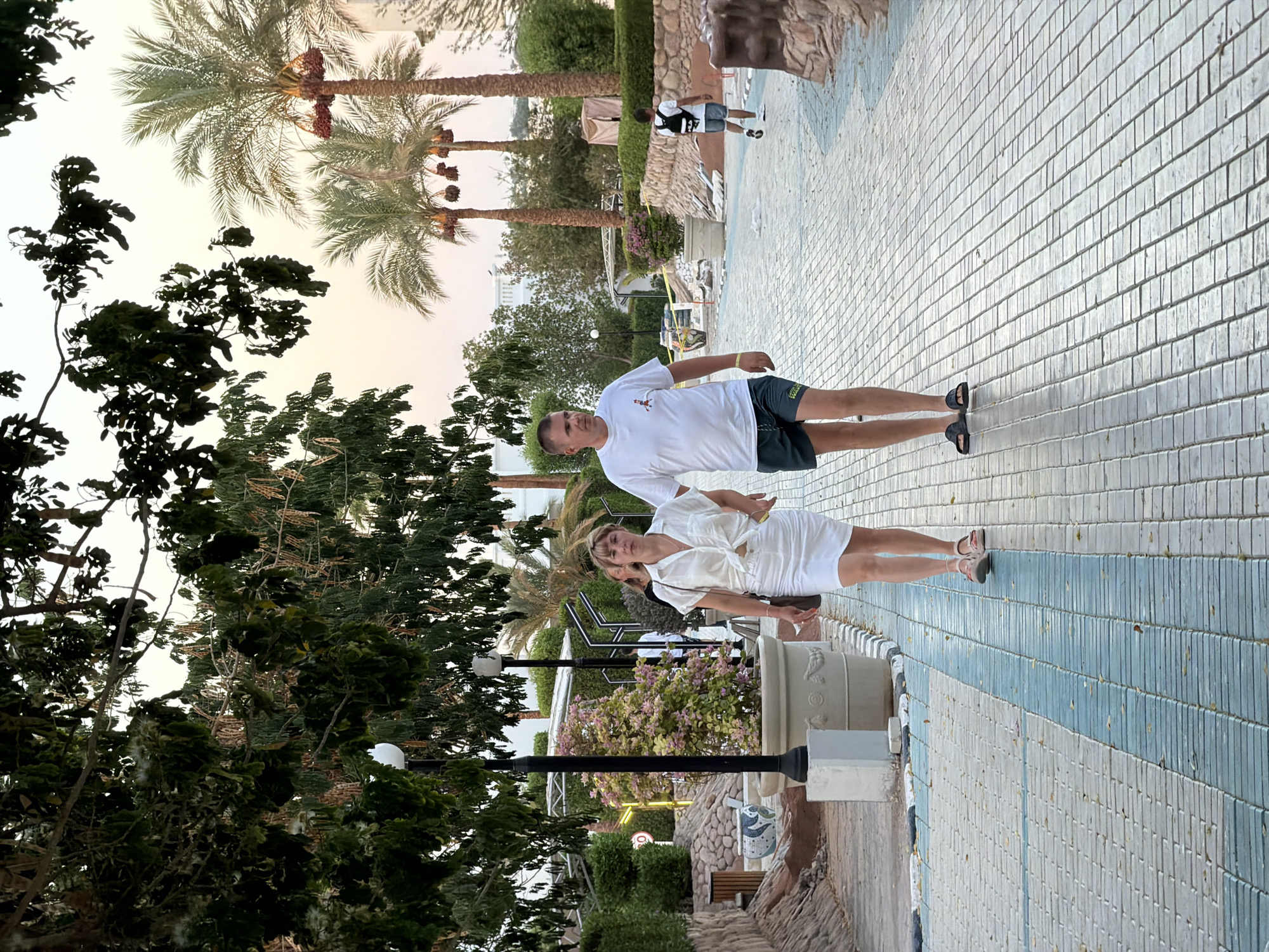 In Sharm El Sheikh, Egypt, a man and a woman are walking hand-in-hand on a paved walkway lined with decorative tiles and large planters. The man, wearing a white t-shirt and dark shorts, walks on the right side of the path. The woman, in a white blouse and skirt, is to his left. In the background, palm trees, lush greenery, and streetlights are visible. A second man, wearing a backpack and shorts, walks in the distance. The sky appears overcast.