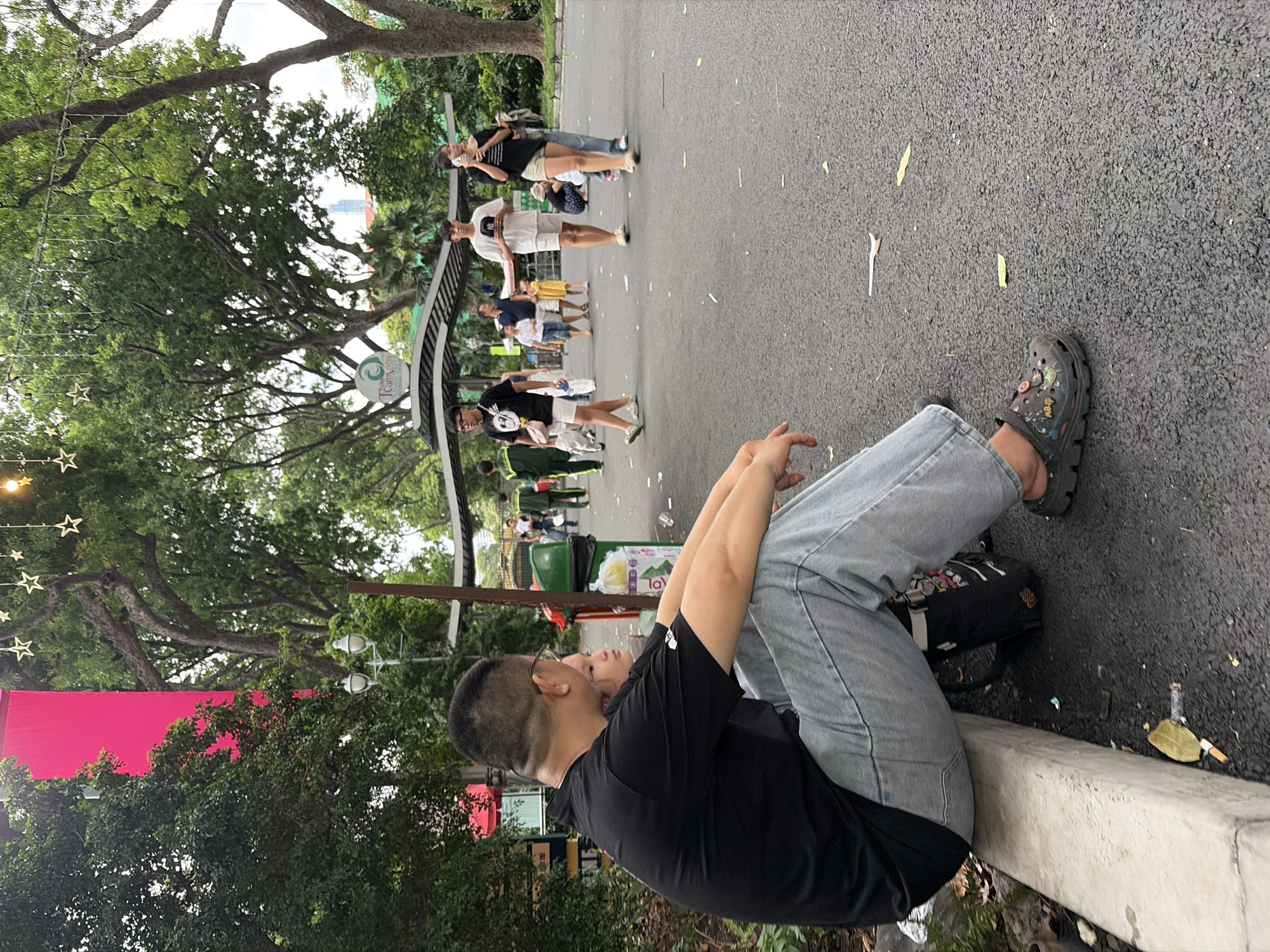 The image depicts a daytime scene within the Saigon Zoo and Botanical Garden (Thảo Cầm Viên Saigon) in Ho Chi Minh City, Vietnam.
In the foreground, a man of East Asian descent, wearing a black t-shirt, light blue jeans, and eyeglasses, sits on a concrete curb on the left. His hands are clasped over his knees, and he wears dark, embellished clogs on his feet. A small black backpack rests beside his left leg. Partially visible behind him is a young child's face, looking forward. The ground in front of them is dark asphalt, scattered with litter including yellow leaves, white debris, and cigarette butts.
In the midground, an asphalt path stretches into the distance, populated by several pedestrians. Individuals include a person in a green uniform walking away, a young man in a black t-shirt and white shorts with a pink shoulder bag walking towards the viewer, another young man in a white t-shirt and shorts holding a cap, and a woman in a black t-shirt and light shorts talking on a mobile phone. A green trash can stands behind the seated man, and a structure with a dark, curved roof, featuring the "Thảo Cầm Viên Saigon" logo, is visible further along the path.
The background is dominated by lush, dense green foliage and tall trees. Through the trees on the far left, a bright pink or red vertical banner is discernible. Overhead, decorative string lights with white star-shaped ornaments and circular bulbs are strung across the scene. Distant urban buildings, including a tall skyscraper, are faintly visible through the trees, indicating the garden's city location. Street lamps are interspersed within the tree line.
