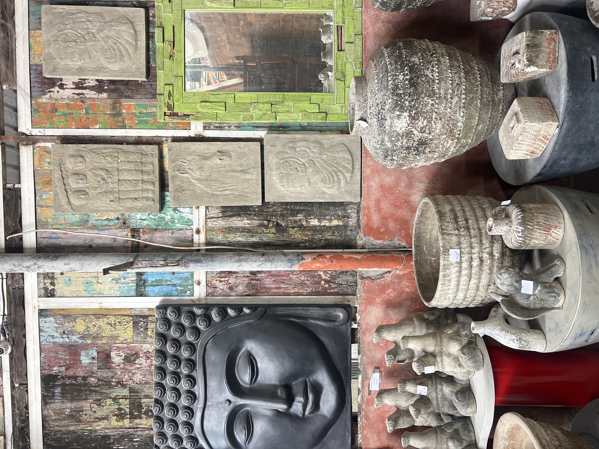 In North Kuta, Indonesia, an eclectic shop displays a variety of decorative items. On the left, a large, dark gray, stylized Buddha head is mounted on a distressed, multi-colored wooden wall. To its right, three stone relief panels are affixed to the same wall: a rectangular panel depicting several figures in a row, a square panel showing a horse, and another rectangular panel with a profile of a person. A bright green, distressed wooden mirror frame sits to the right of these panels, reflecting a cluttered interior. In the foreground, an assortment of ceramic and stone objects are arranged on red and gray pedestals. These include several small, light gray animal-like figurines, a large, weathered, textured pot, and smaller vases and decorative blocks. The overall impression is one of a curated collection of art and decor, likely sold in a market or specialized shop.
