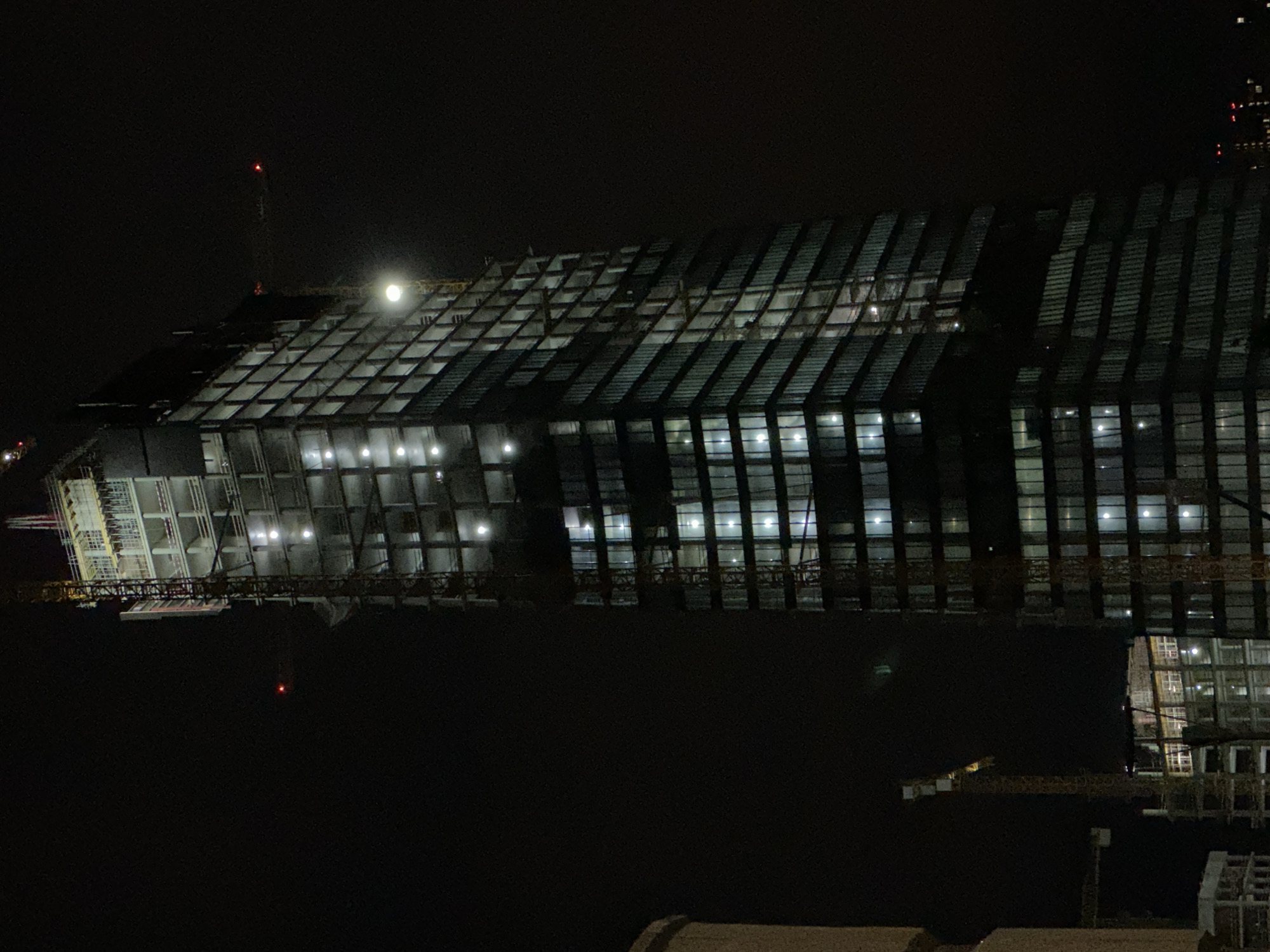 A nighttime, low-angle, medium shot captures the skeletal structure of a tall skyscraper under construction in an urban setting. The building, mostly unclad, features rows of concrete floors and external scaffolding. Lights are visible from within the building, illuminating some of the unfinished floors. The skyscraper is flanked by construction cranes, some with red lights indicating their presence. The surrounding environment is dark, suggesting the image was taken at night. The overall impression is one of a massive, ongoing construction project dominating the nocturnal cityscape.