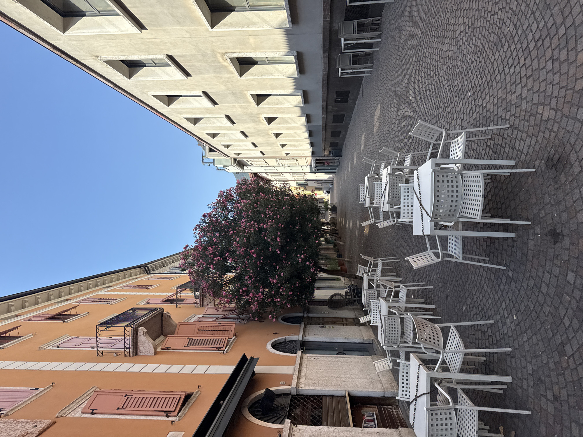 This image depicts a street scene in Trento, Italy, likely in the historic center, characterized by its cobblestone pavement and architecture. The street is lined with buildings on both sides. The building on the left features an orange facade with pink shutters and a wrought-iron balcony. There are arched entrances and windows on the ground floor. A large tree with pink blossoms is prominently situated between the two buildings, adding a touch of color to the scene.
On the street, numerous white metal tables and chairs are arranged, suggesting an outdoor seating area for a cafe or restaurant. The tables are mostly square, and the chairs have a distinctive grid pattern on their backs and seats. Some tables have chains draped across them, possibly for security or to keep them together. The cobblestone pavement is laid in a circular pattern in the foreground, which gradually transitions to a more uniform arrangement further down the street.
The building on the right has a light beige facade with numerous rectangular windows that are recessed. The shadows cast by the window frames create a repeating geometric pattern on the wall. The sky above is clear and blue. No people are visible in the scene. The overall impression is one of a quiet, early morning or late afternoon in a European city, with the focus on the architectural details and the arrangement of the outdoor seating.
