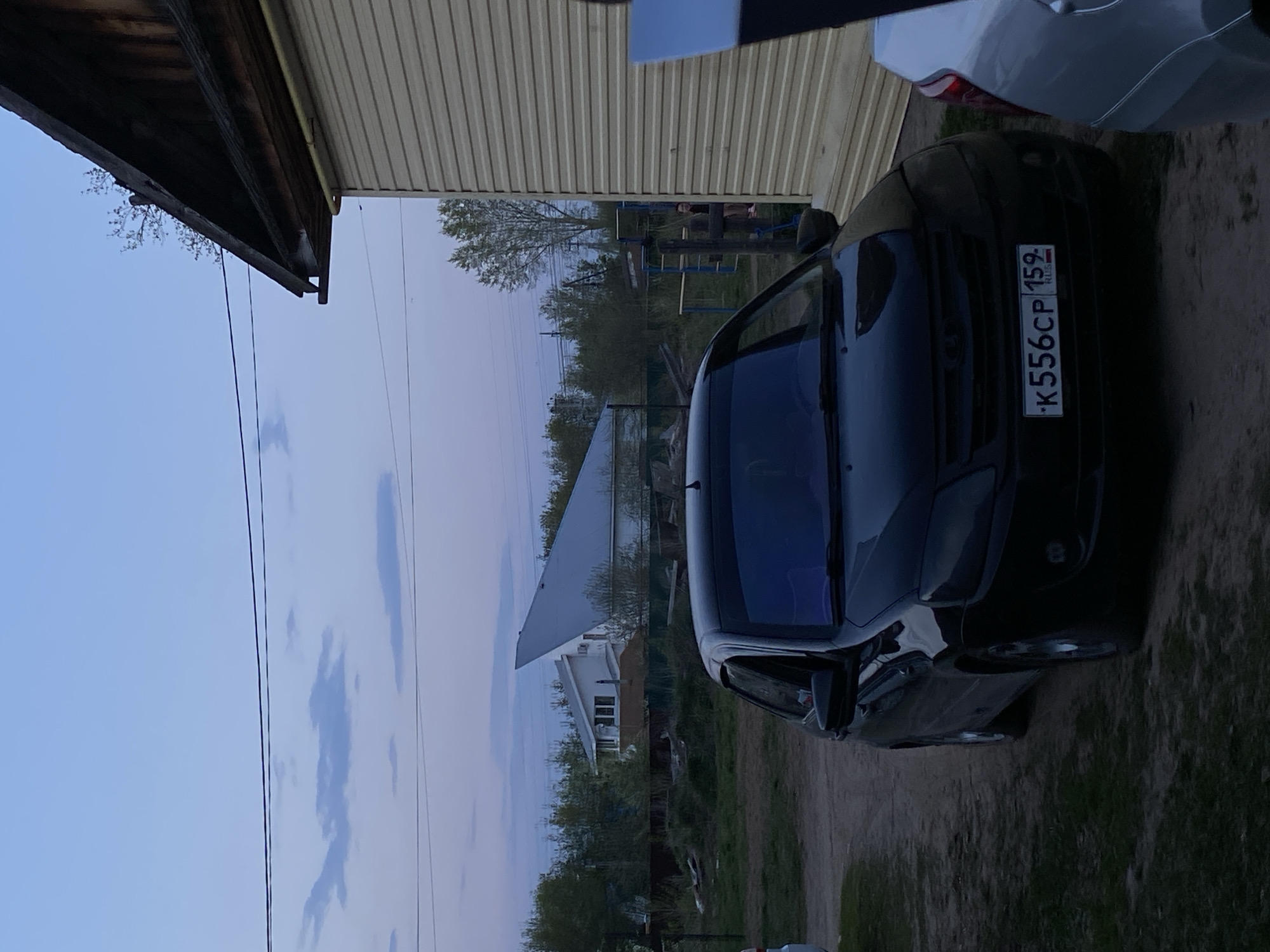 The image shows a black car parked near a building with light-colored siding. The license plate of the car is visible, reading "K556CP 159 RUS," indicating registration in Russia. In the background, a residential area with houses and trees can be seen under a cloudy sky. The location appears to be in a suburban or rural setting. This is in Chaikovsky, Russia. A swing set is visible in the background.