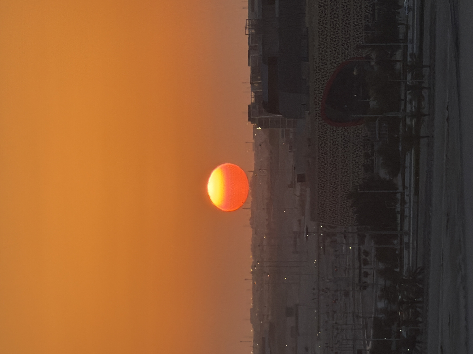 An image captures a sunset over a city. The sun, a bright orange orb with a yellow center, is prominent in the sky, positioned slightly above the horizon. The sky is a gradient of warm orange tones. In the midground, a low-lying cityscape is visible, with silhouetted buildings and structures, some with illuminated windows or lights. The foreground reveals more urban elements, including streetlights and trees. The overall scene depicts a dusk or dawn setting in an urban environment.