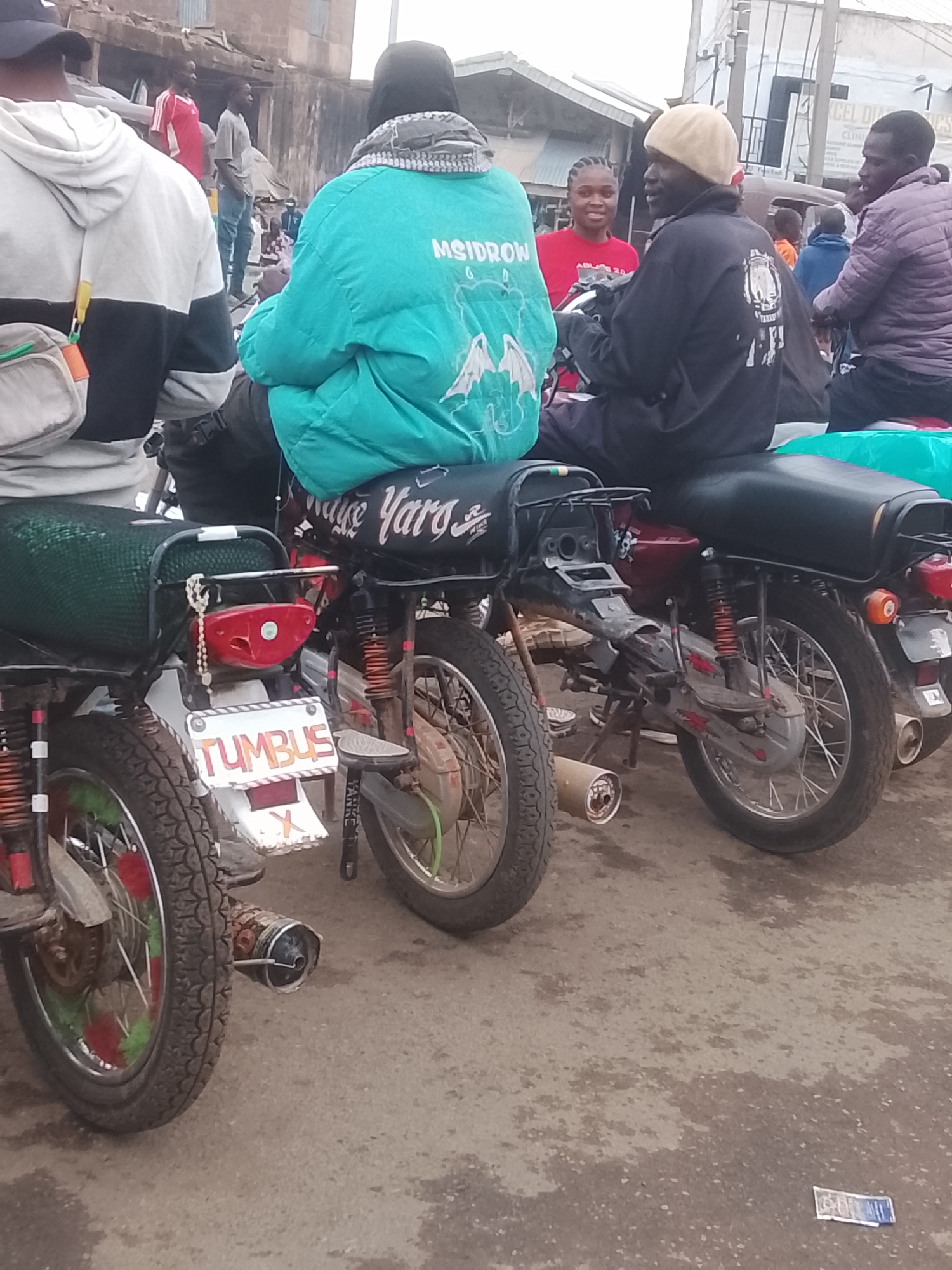 The image depicts a scene in what appears to be Gwong, Nigeria. Three motorcycles are parked in a row on a paved surface that shows signs of wear and discoloration. People are seated on the motorcycles, facing forward. The motorcycle furthest to the left has a license plate that reads "TUMBUS." The tire of this motorcycle features a red and green design. The person on the leftmost motorcycle is wearing a hooded sweatshirt and is positioned with their back to the camera. To their right, the person on the second motorcycle is wearing a bright turquoise jacket with white lettering and designs, as well as black trousers. "Yaro" is written in cursive across the bike's fender. The third person, on the rightmost motorcycle, wears a black jacket with a graphic design on the back and a beige cap. Another person wearing red is visible standing between them. People are also seen standing in the background, indicating a populated environment. Litter, including a bill of currency, is visible on the ground near the motorcycles. Buildings and other structures are partially visible in the background.