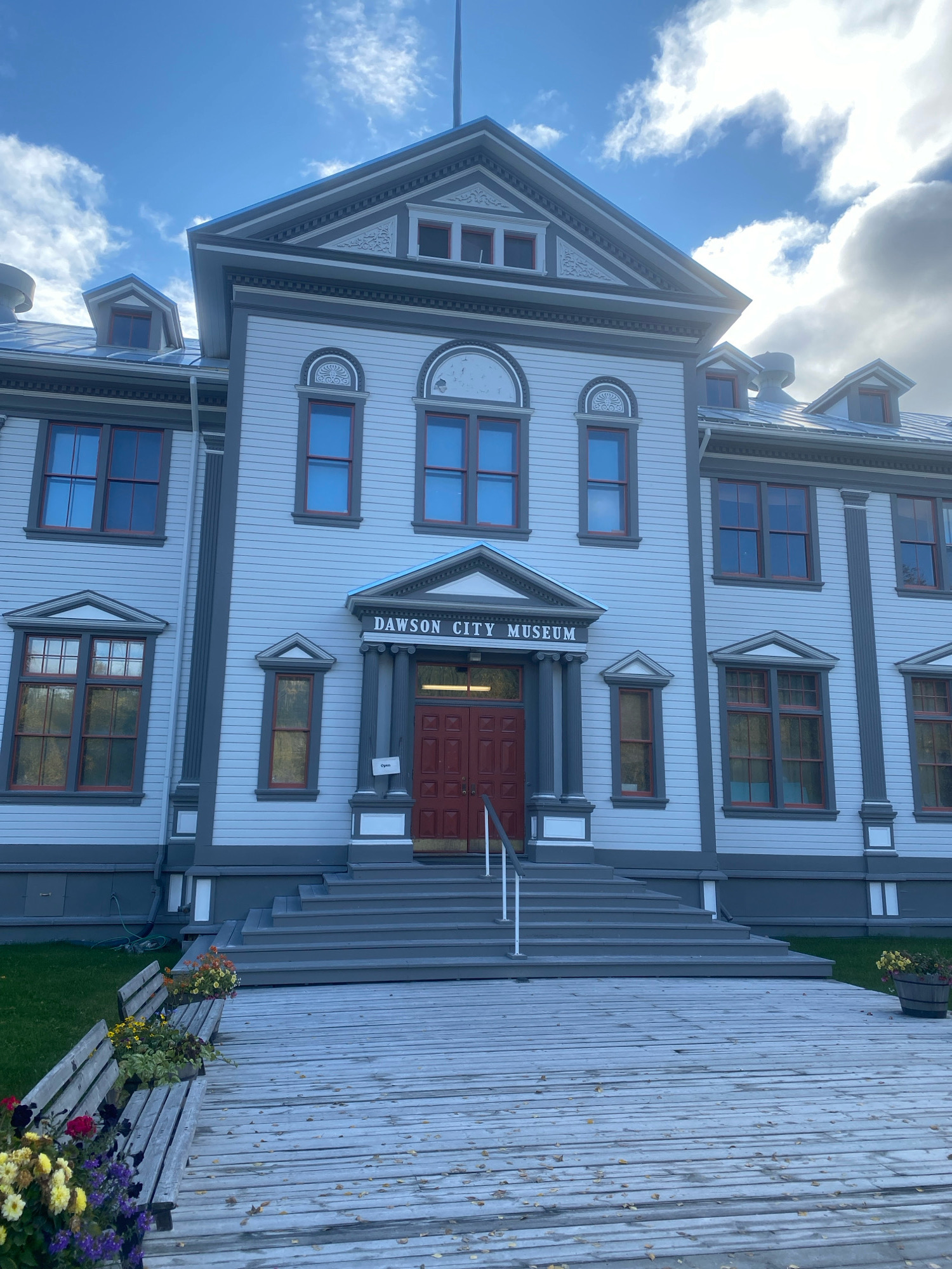 The image depicts the Dawson City Museum, a historic building located in Dawson City, Yukon, Canada. The museum is a two-story structure with a light blue facade and dark grey trim. It features a prominent central entrance with a pedimented portico supported by columns. The word "DAWSON CITY MUSEUM" is inscribed above the entrance. Red double doors are visible within the portico.
A wide, wooden deck leads up to a set of grey stairs that ascend to the entrance. To the left of the deck, two wooden benches are situated, adorned with pots of colorful flowers. The deck is covered with fallen autumn leaves. Grass areas surround the building and the deck.
The sky above is partly cloudy with patches of blue. The architecture of the building suggests a historical era, common in Dawson City due to its Gold Rush history. No people are visible in the image. The scene is captured on a clear day, likely in the fall given the foliage.