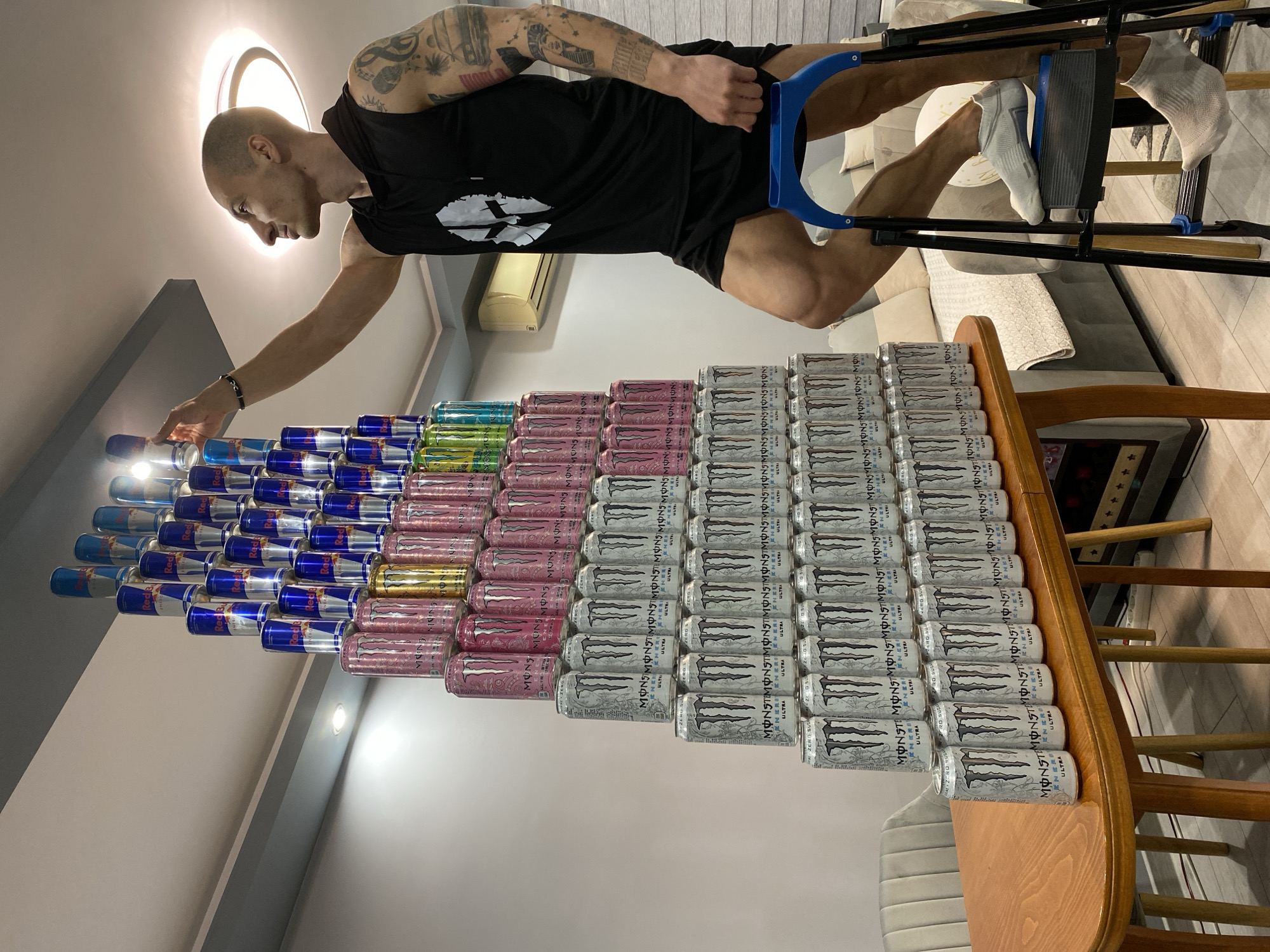 A man with tattoos on his arm stands next to a table stacked with multiple rows of Red Bull and Monster energy drinks, constructing a pyramid shape. He is using crutches. The environment appears to be an indoor living space. The location, Pleven, Bulgaria, cannot be confirmed from the image.