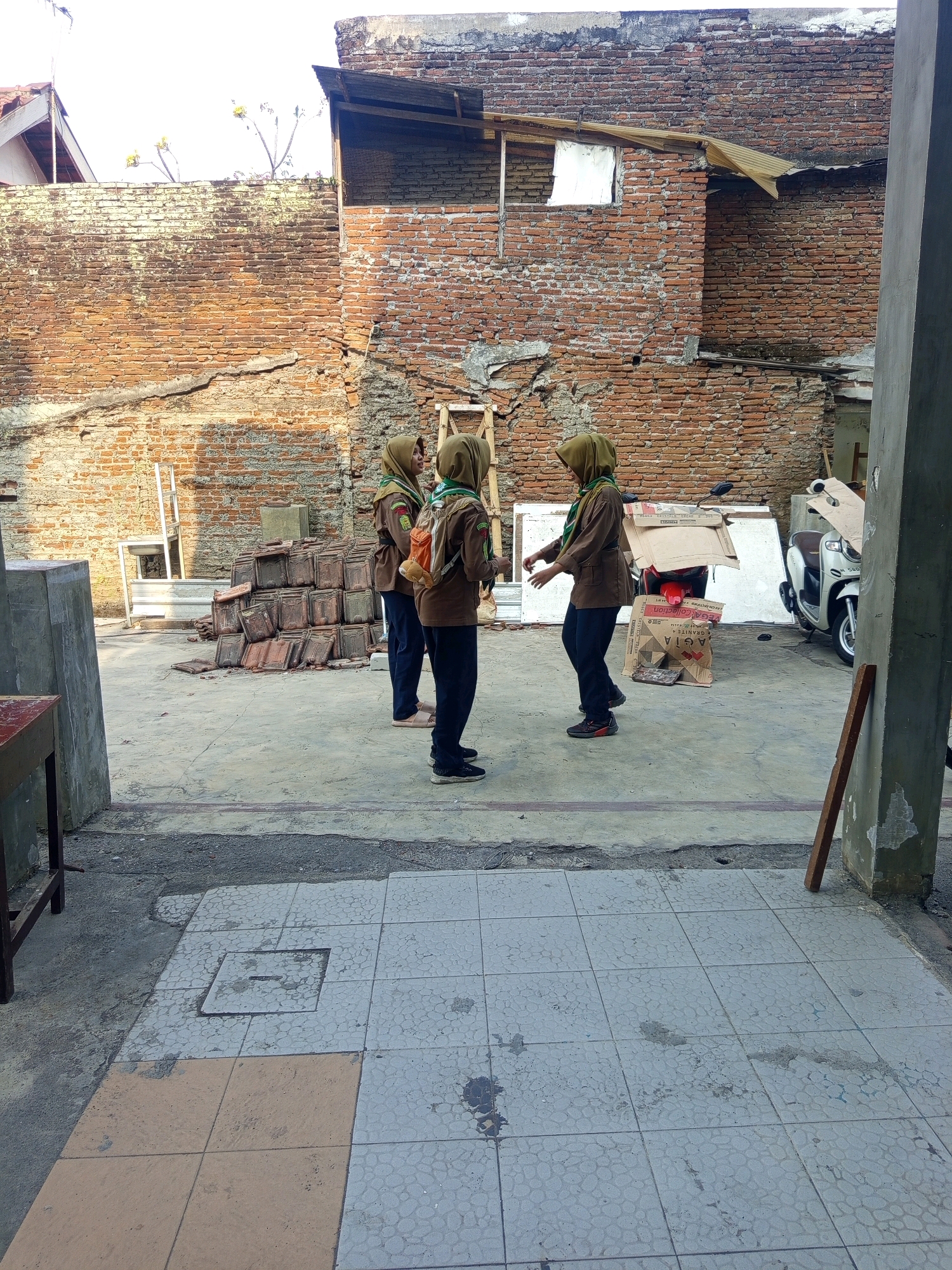 Three young women in matching brown uniforms and green headscarves are standing in an outdoor courtyard. The courtyard is paved with concrete and tiled areas. Behind the women is a brick wall, partially covered with moss and damage, with several stacks of roof tiles leaning against it. A wooden ladder is also visible against the wall. In the background, there is a partially constructed or dilapidated building with a corrugated metal roof. To the right, a white scooter is parked next to a large cardboard box. The overall environment appears to be an urban setting in Malang, Indonesia, possibly a schoolyard or a residential area under renovation.