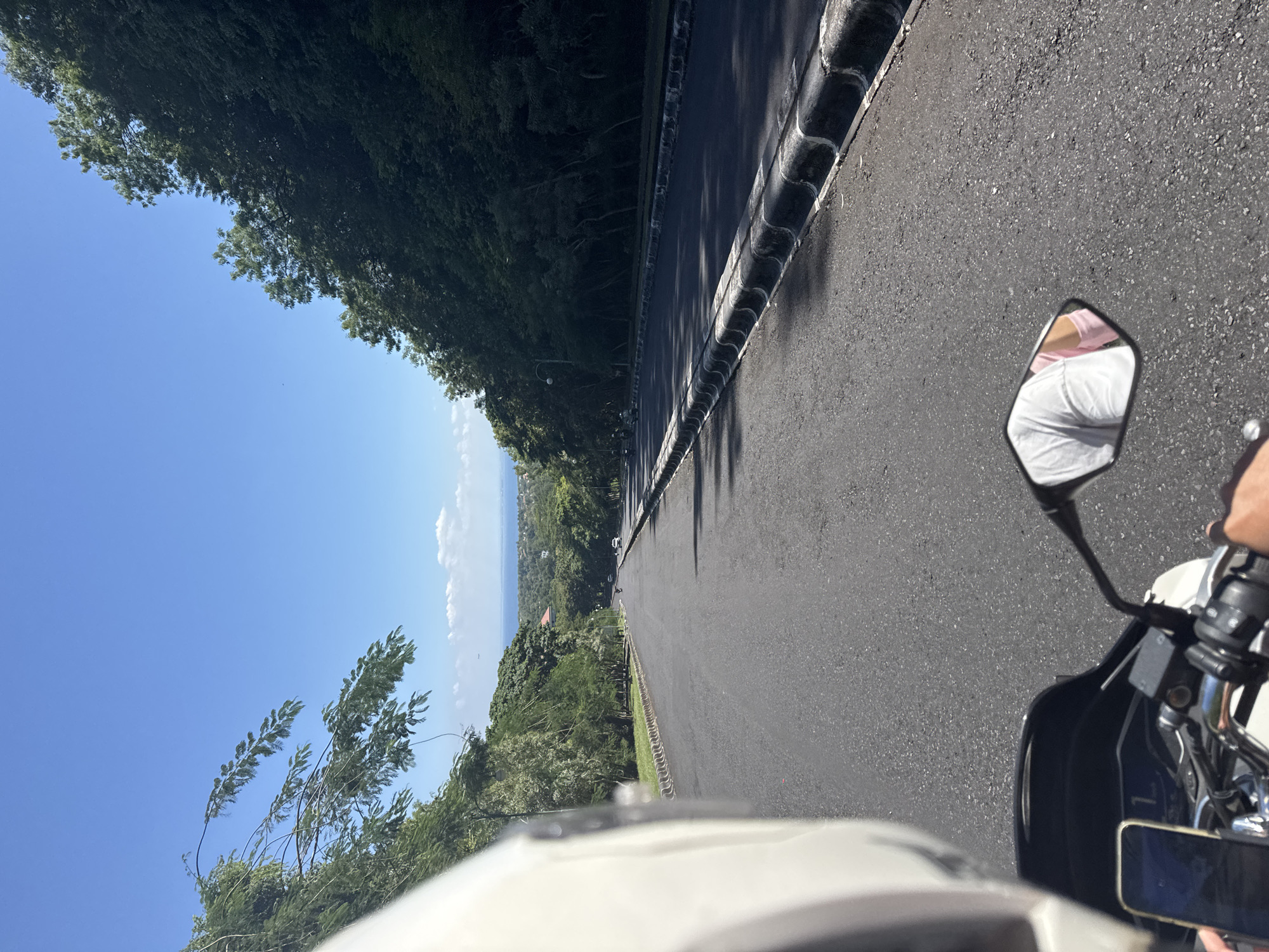 A low-angle, tilted perspective shows an asphalt road with a textured, black safety barrier lining the right side. The road appears to descend towards a visible ocean horizon under a clear blue sky, with sporadic cloud formations near the distant water. Dense green trees and foliage border the road on both sides. The front of a white motorcycle and its side mirror, reflecting a person wearing a white shirt and pink undershirt, are in the foreground. The location is potentially in the city of Unknown, Unknown.