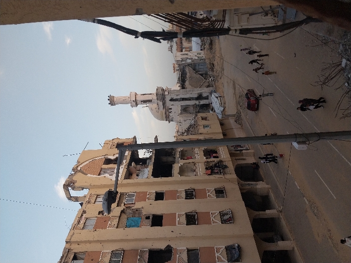 A damaged building with missing walls and windows stands beside a partially destroyed mosque with a distinct minaret. Debris litters the ground and surrounding area. Figures walk along the street, where a red car is parked. Electrical wires stretch across the top of the frame, adding to the sense of disarray and urban decay. The overall scene conveys a sense of destruction and the aftermath of a conflict.