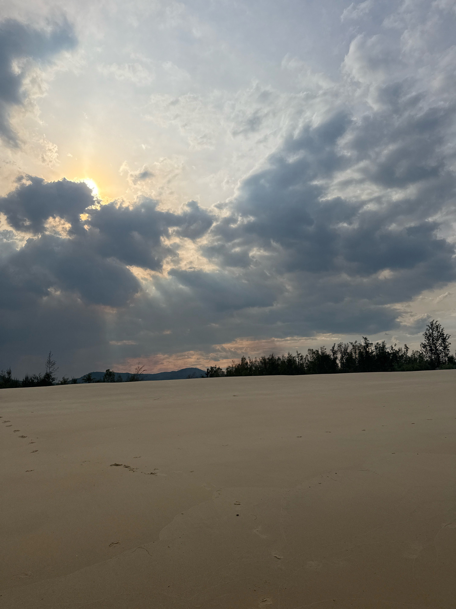 A landscape photograph captures a serene view of a sand dune against a dramatic sky. The foreground is dominated by the vast expanse of a sandy desert, with faint footprints trailing across the surface, suggesting recent passage. In the middle ground, a dense line of trees forms a dark silhouette against the horizon, with a range of hills visible behind them. The sky above is a spectacle of light and shadow, with heavy, dark clouds partially obscuring a bright, sunlit area where the sun's rays pierce through, creating a dramatic, almost divine illumination. The overall impression is one of natural beauty and a touch of mystery, evoking a sense of solitude and contemplation.