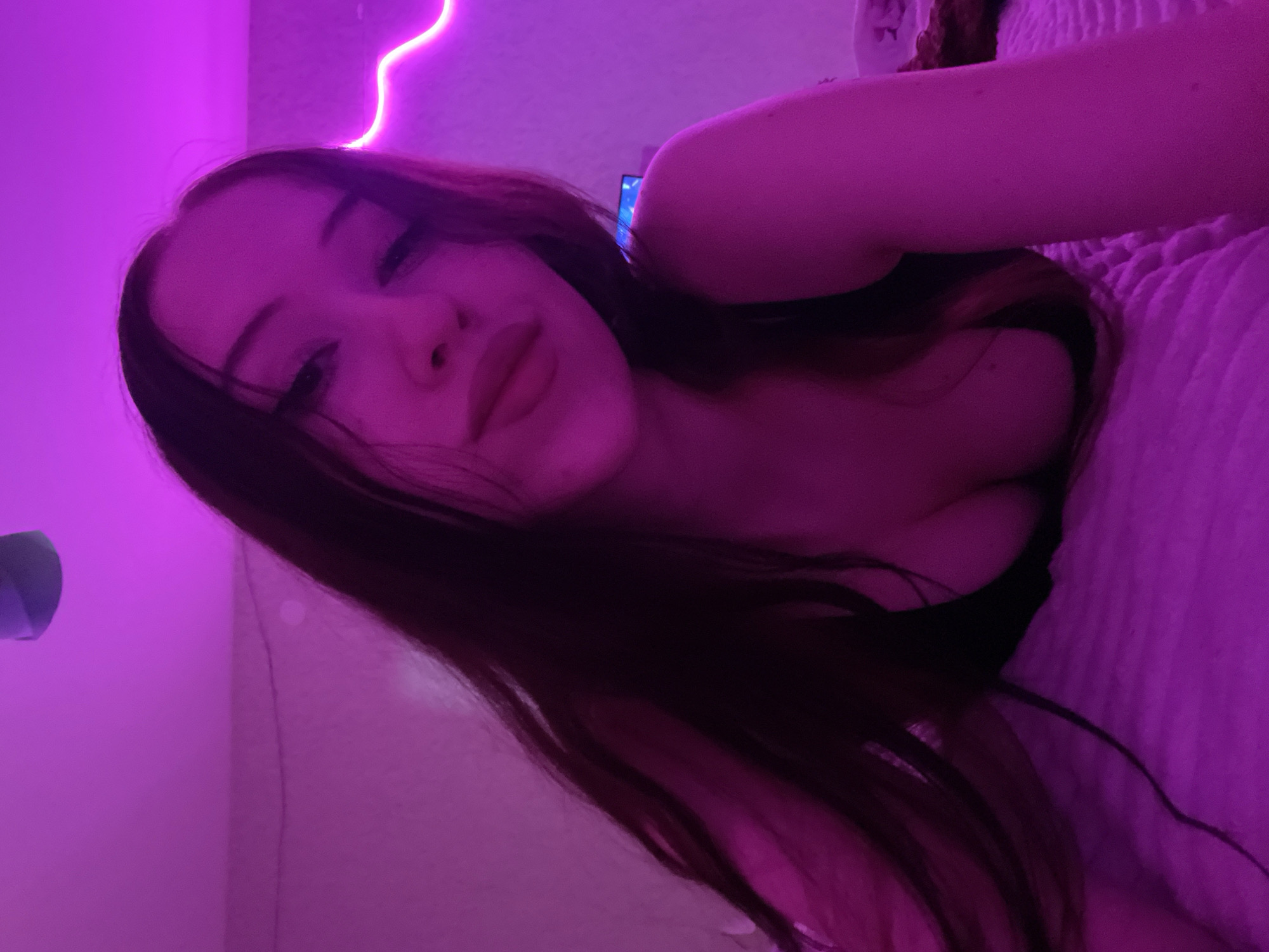 The image shows a young woman with long, dark hair, lying down on a textured surface, possibly a bed. She is wearing a dark top that reveals her cleavage. The lighting is dominated by a vibrant pink or purple neon glow, likely from LED strip lights. The background appears to be a wall with some subtle texture and a faint outline of what might be a ceiling fan or light fixture. There is also a hint of a television screen in the upper right background, casting a blueish light. The overall atmosphere is intimate and dimly lit, with the neon lights creating a moody ambiance. There is no discernible connection to Votkinsk, Russia.
