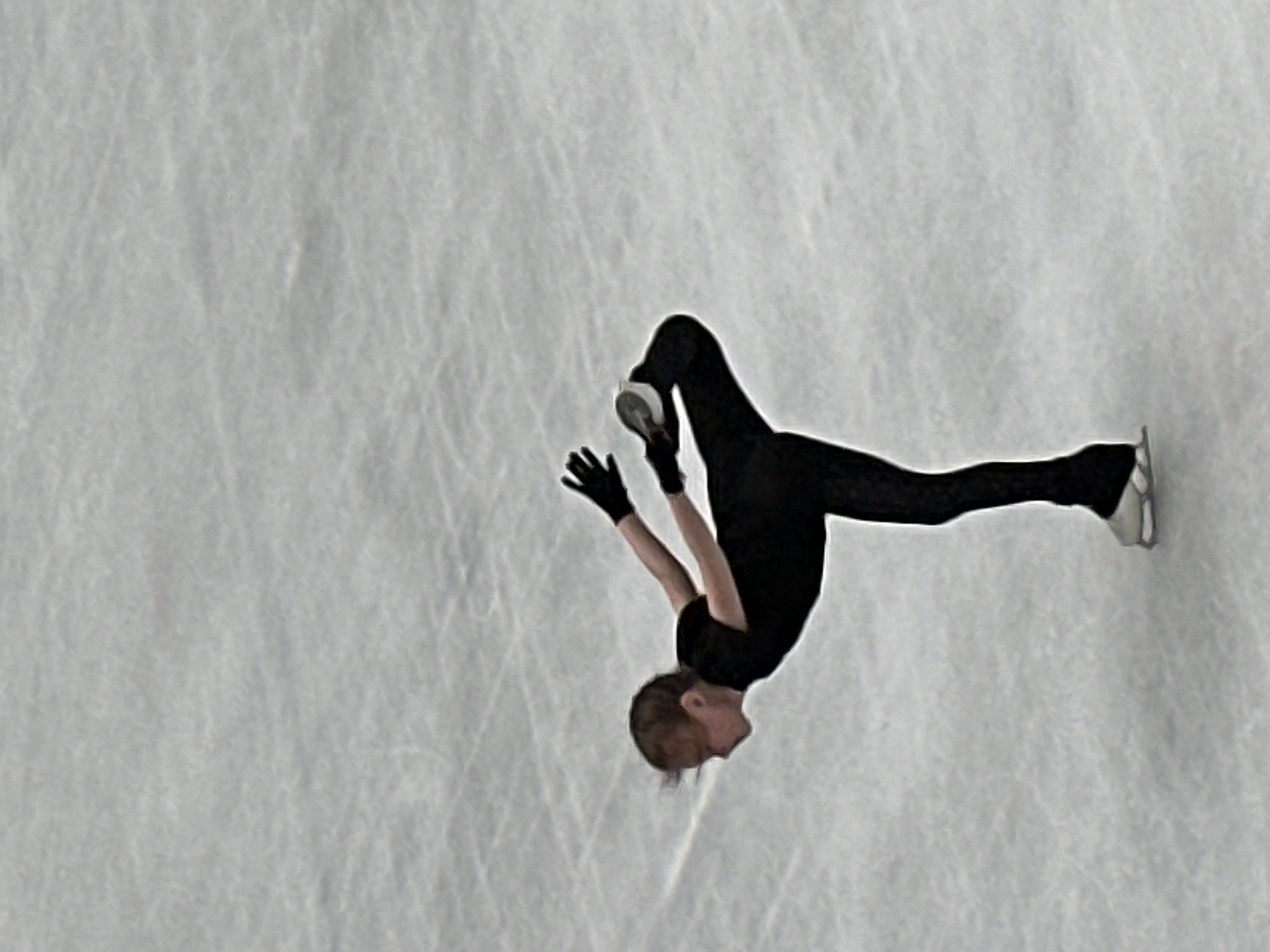 A figure skater in a black outfit is performing a Biellmann spin on an ice rink. The skater is facing downwards with their head close to their raised leg. The image is taken from above, showing the texture of the ice. The location, Nizhny Novgorod, Russia, is not discernible from the image.