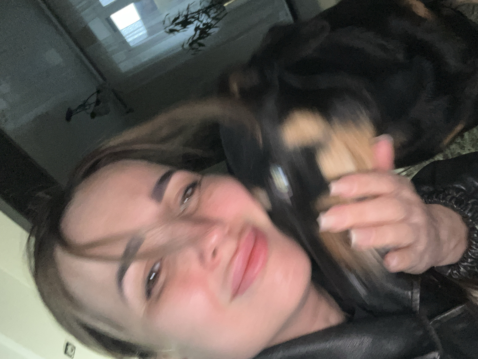 A selfie-style photograph shows a woman with light skin and dark eyebrows holding a dog with black and tan fur. The woman is positioned in the lower-left corner of the frame, with her face slightly blurred. She has dark hair with lighter streaks. Her mouth is closed in a slightly pursed expression. The dog occupies the upper-right portion of the image, and the woman's hand is visible near its head. The environment appears to be indoors. A window is visible in the upper-left, showing light. A dark, leafy plant is visible in the window.
The location is Unknown, Unknown.