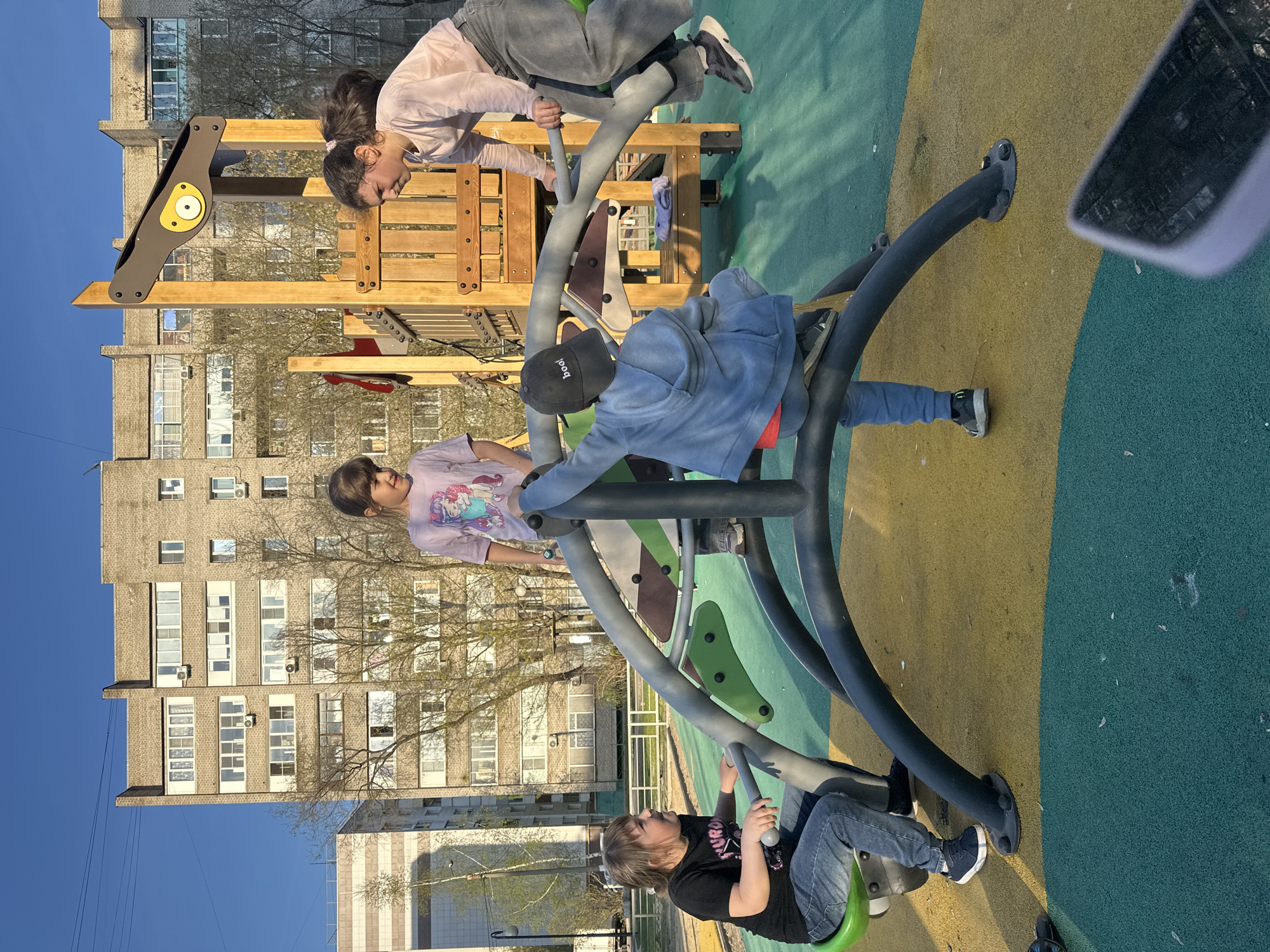 The image captures a playground scene in Tomilino, Russia. Four children are actively engaged on playground equipment. One child is positioned at the top of a wooden climbing structure with a yellow panel, while three others are interacting with a rotating, multi-seat piece of equipment made of metal. The children are dressed in casual clothing. Behind the playground, a tall, multi-story apartment building is visible. The playground itself is constructed with a padded surface of green and yellow. The sky is blue and the weather is sunny.
