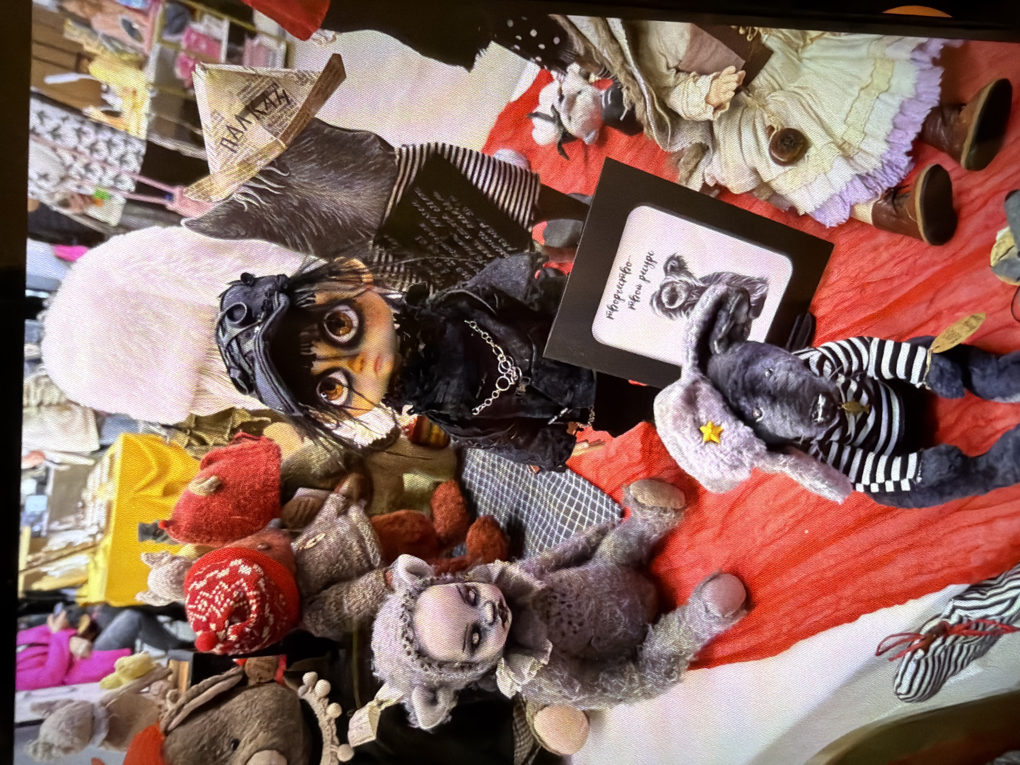The image depicts a collection of dolls and stuffed animals arranged on a surface that appears to be a blanket or fabric with red and white sections. The scene includes a doll with large eyes and a black outfit, a stuffed rabbit with a star on its ear, and a small framed picture with text in Russian, which reads "творчество новой природы" ("creativity of new nature"). Other items include various knitted hats, a doll in a dress with brown boots, and a stuffed animal resembling a bear or sheep.
Without further evidence, it is impossible to conclusively confirm the location in Moscow, Russia. The Russian text hints at a possible Russian association, but it is insufficient for definitive localization.