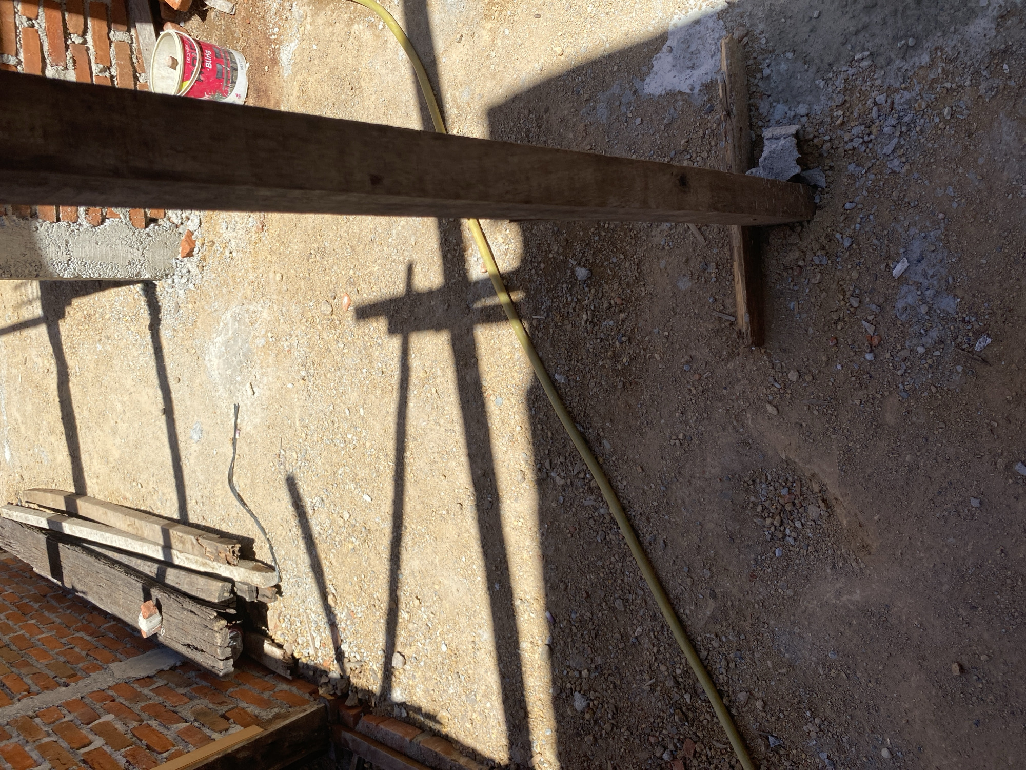 A construction site under a bright sun. A pile of old wooden beams leans against a brick wall on the left. A yellow hose snakes across the sandy, gravelly ground. In the foreground, a thick wooden post stands vertically, casting a long shadow. Nearby, a red and white bucket sits on the ground. The scene is littered with small rocks and debris, suggesting ongoing construction work.