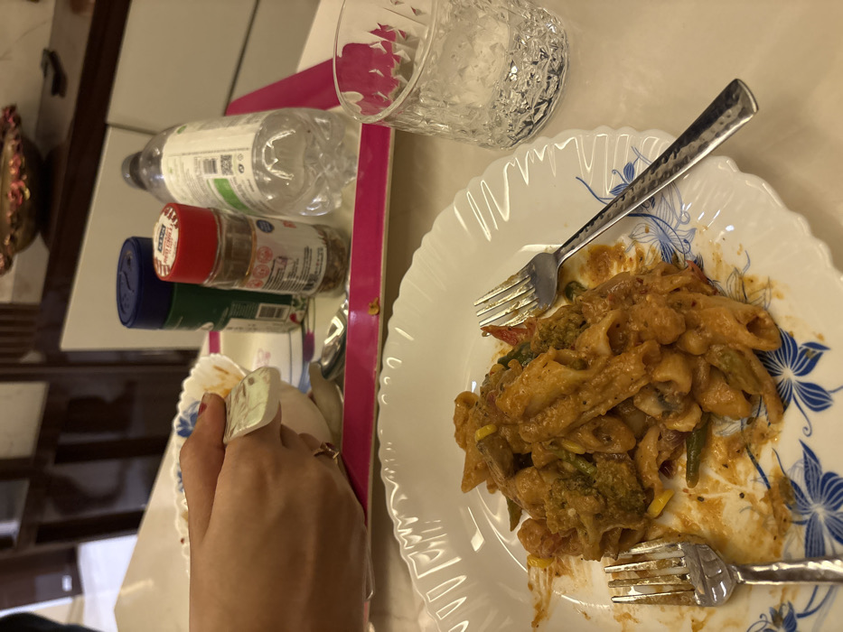 A plate of penne pasta with vegetables, covered in a creamy orange sauce, sits on a table. Two forks are placed on the plate. Next to the plate on a pink tray are a glass of water, a bottle of water, and three spice containers. A hand holding a white napkin is also visible.