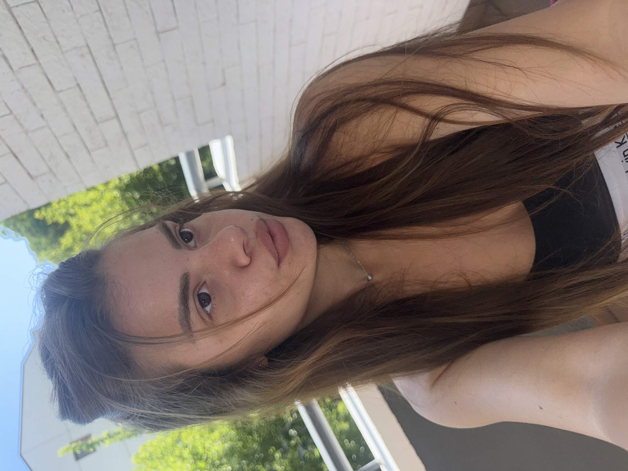 A young woman with long, brown hair pulled up in a bun is standing on a balcony. She is wearing a black top and a silver necklace. The background shows green trees, a white brick wall, and a blue sky. The scene appears to be a sunny day in Plovdiv, Bulgaria.