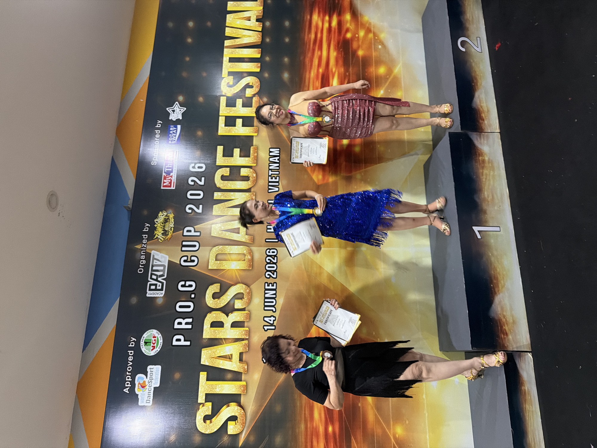 Three women are standing on a podium in front of a backdrop that reads "PRO.G CUP 2026 STARS DANCE FESTIVAL" and "14 JUNE 2026 | HANOI VIETNAM". The woman on the left is wearing a black dress with fringe and leopard print heels. She is holding a certificate and a medal. The woman in the middle is wearing a sparkly blue dress and gold heels. She is holding a certificate. The woman on the right is wearing a sparkly pink dress and gold heels. She is also holding a certificate and a medal. The podium has the numbers 1 and 2 visible on it. The backdrop also features logos of various organizations, including "Approved by World DanceSport Federation" and "Organized by GRUZ" and "Sponsored by My Thanh Anh and Pocari Sweat". The scene appears to be an award ceremony for a dance competition.