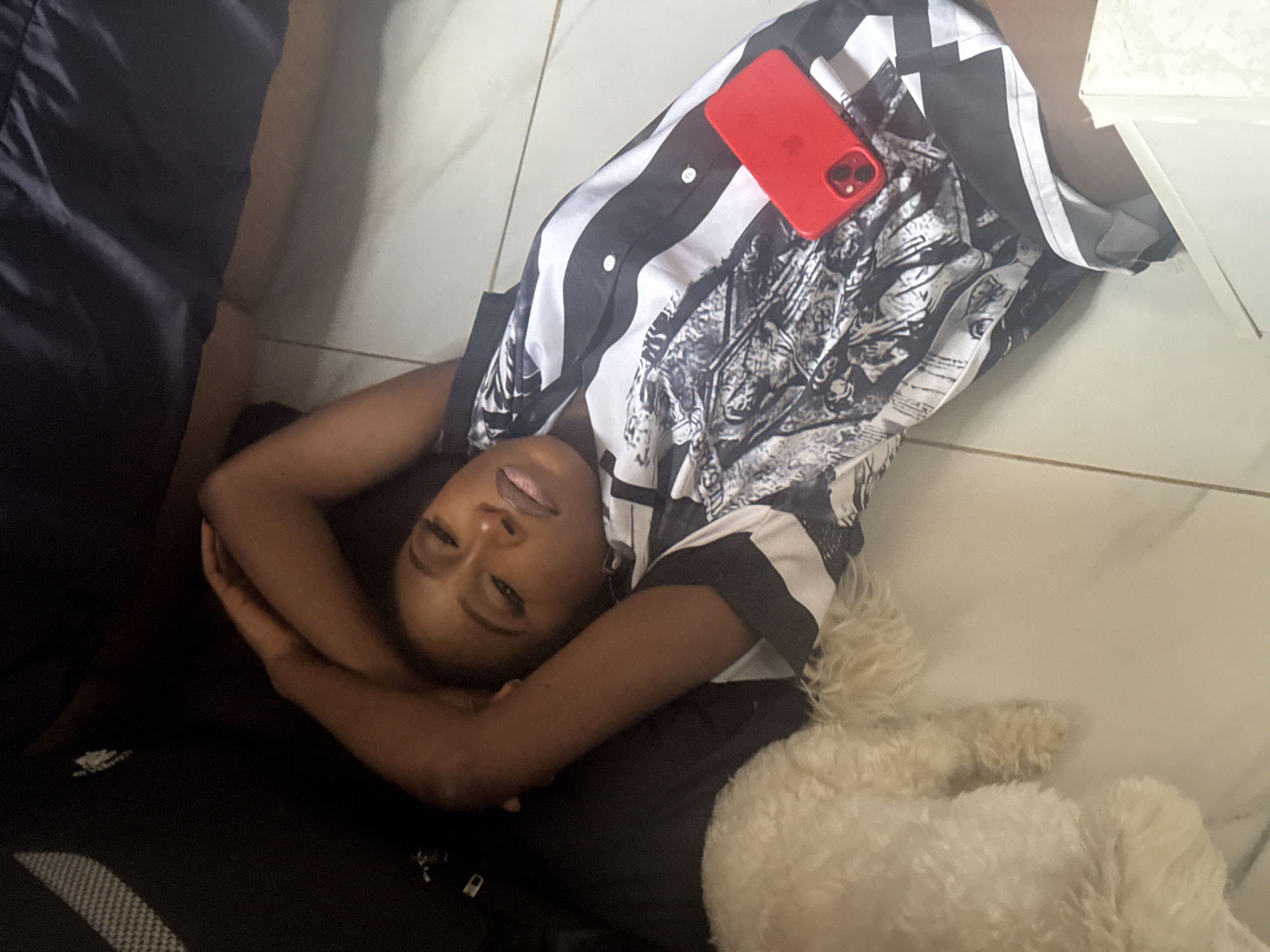 A Black woman is lying on her back on a tiled floor, her arms crossed over her head. She is wearing a black and white striped shirt with a black and white graphic print on the front. A red cell phone is placed on her chest, with its camera lens facing upwards. To her left, a small, fluffy white dog is lying on the floor. The background is dark, with hints of furniture. The lighting suggests an indoor setting, possibly a residential apartment. The image does not provide any specific information that would indicate the location as Abuja, Nigeria.