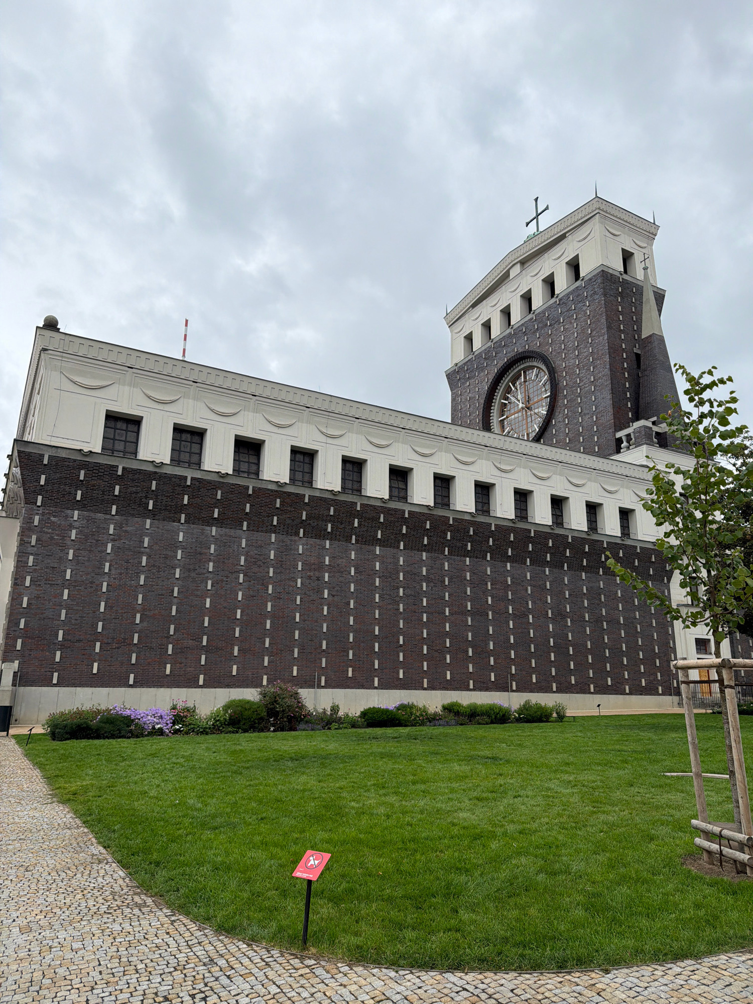 The image shows the Church of the Most Sacred Heart of Our Lord, a Roman Catholic basilica located in the Vinohrady district of Prague, Czechia. The church is characterized by its distinctive modern architecture, featuring a tall, square tower topped with a cross. The facade is primarily constructed of dark brick with lighter horizontal bands and a prominent circular window. In the foreground, there is a well-maintained lawn and a cobblestone pathway. A small red sign with a white symbol is placed on the grass. The sky is overcast, suggesting a cloudy day.