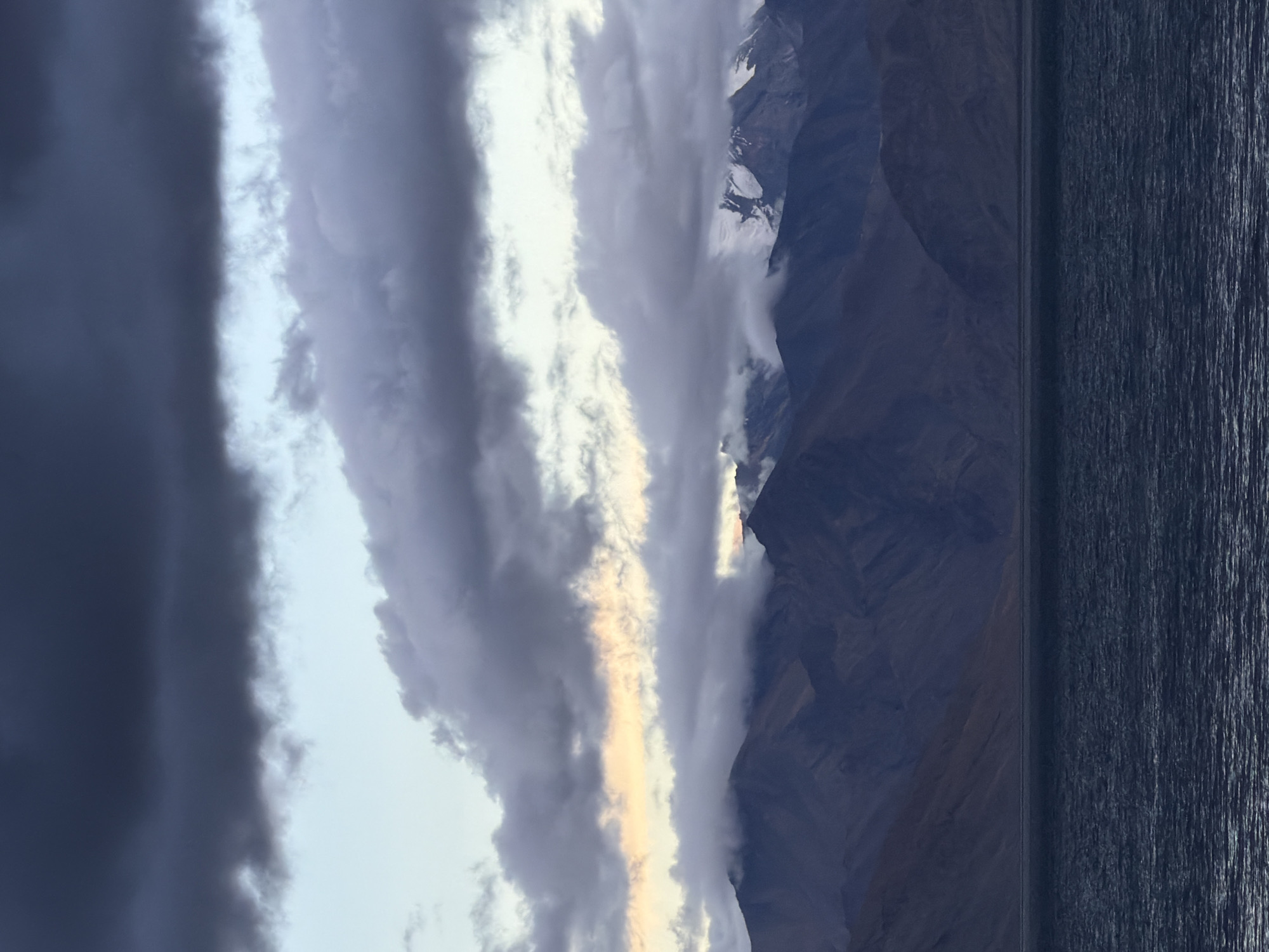This image captures a panoramic view of the Tso Moriri lake in Karzok, India. The scene is dominated by a vast expanse of dark, choppy water in the foreground, reflecting the moody sky above. In the midground, majestic, rugged mountains rise, their slopes appearing a deep, shadowy hue. The peaks are partially shrouded in thick, dramatic clouds that are tinged with soft yellow and orange light, suggesting either sunrise or sunset. Some of the highest mountain peaks are dusted with snow, contrasting with the darker rock formations. The sky is filled with layers of dark, heavy clouds, creating a sense of impending weather or dramatic atmospheric conditions. There are no people or man-made objects visible in the frame, emphasizing the raw, natural beauty of the landscape.