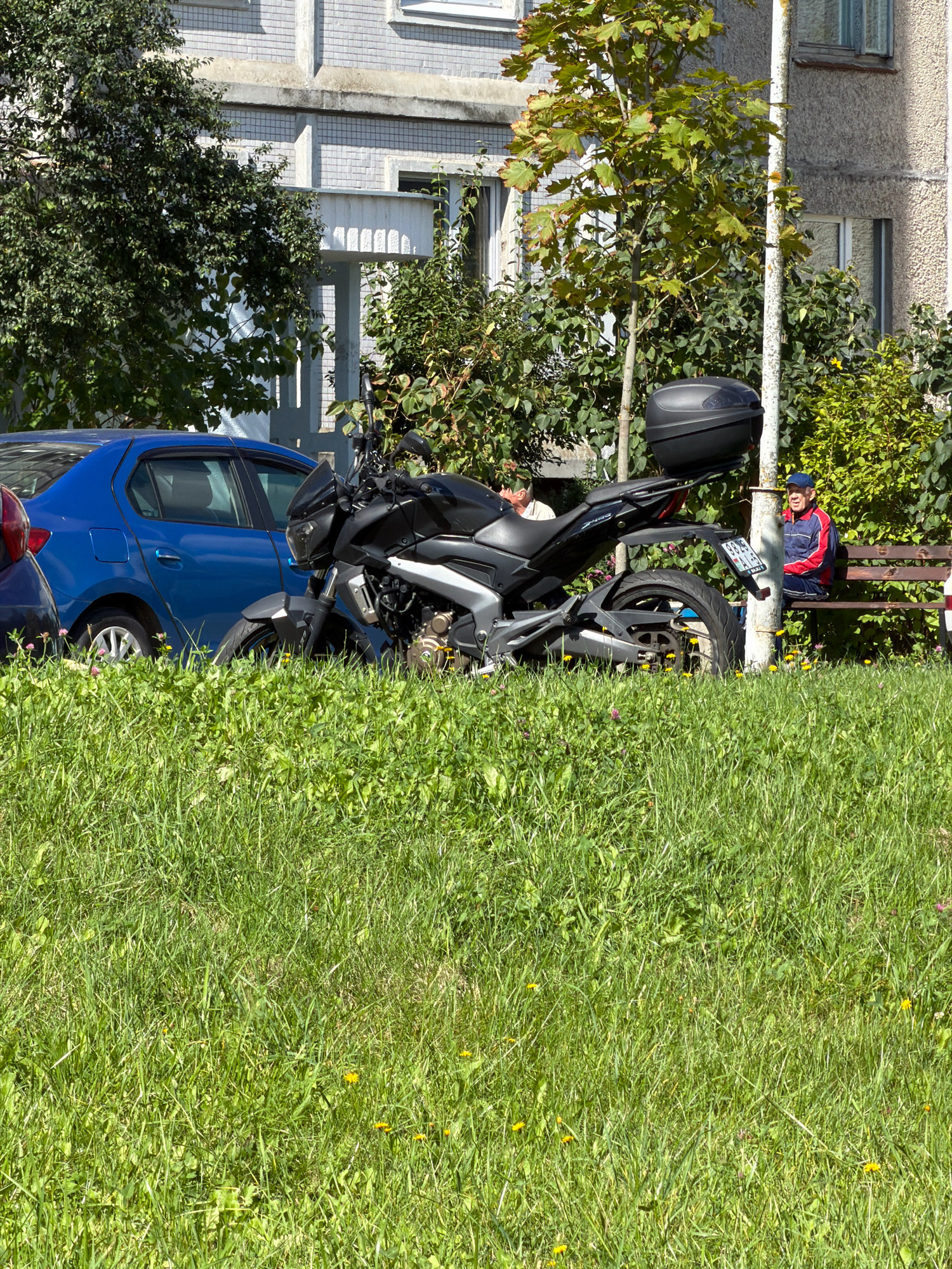 In a sunlit outdoor scene, a black motorcycle is parked in front of a blue sedan, positioned on a patch of green grass. Behind the vehicles, a building with light-colored, textured walls and white window frames stands under a clear sky. Trees with lush green foliage are scattered throughout the environment. To the right, an elderly man wearing a red and blue jacket and a blue cap is seated on a wooden bench, facing the camera and looking towards the motorcycle. A white pole, possibly a lamppost, stands vertically between the motorcycle and the bench. The overall atmosphere suggests a casual outdoor setting within a city.