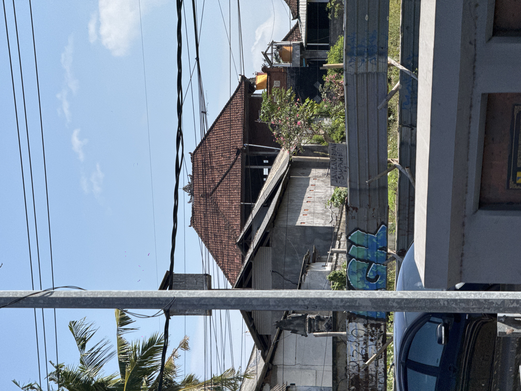 The image shows a street scene in Sukawati, Indonesia, on a bright, sunny day. The perspective is skewed, as if the camera is tilted. In the foreground, a metal railing runs horizontally across the frame, obscuring the lower portion of the scene. Behind it, glimpses of a dark blue vehicle and more metal structures are visible.
The mid-ground reveals a cluster of buildings with traditional tiled roofs, some appearing under construction or in disrepair. Wires crisscross the sky above, strung between poles. A lush green tree with pink flowers adds a touch of nature to the urban landscape. A sign with text is affixed to a vertical metal post.
Further back, more buildings are visible, along with vegetation. The sky is clear blue with a few wispy clouds, suggesting it is midday or early afternoon. The overall impression is of a busy, somewhat weathered, but still vibrant tropical locale. The visible text on the sign reads "JANGAN BUANG SAMPAH DI SINI," which translates to "Do not litter here" in Indonesian, indicating a public space. The graffiti on a metal surface adds a touch of urban art.