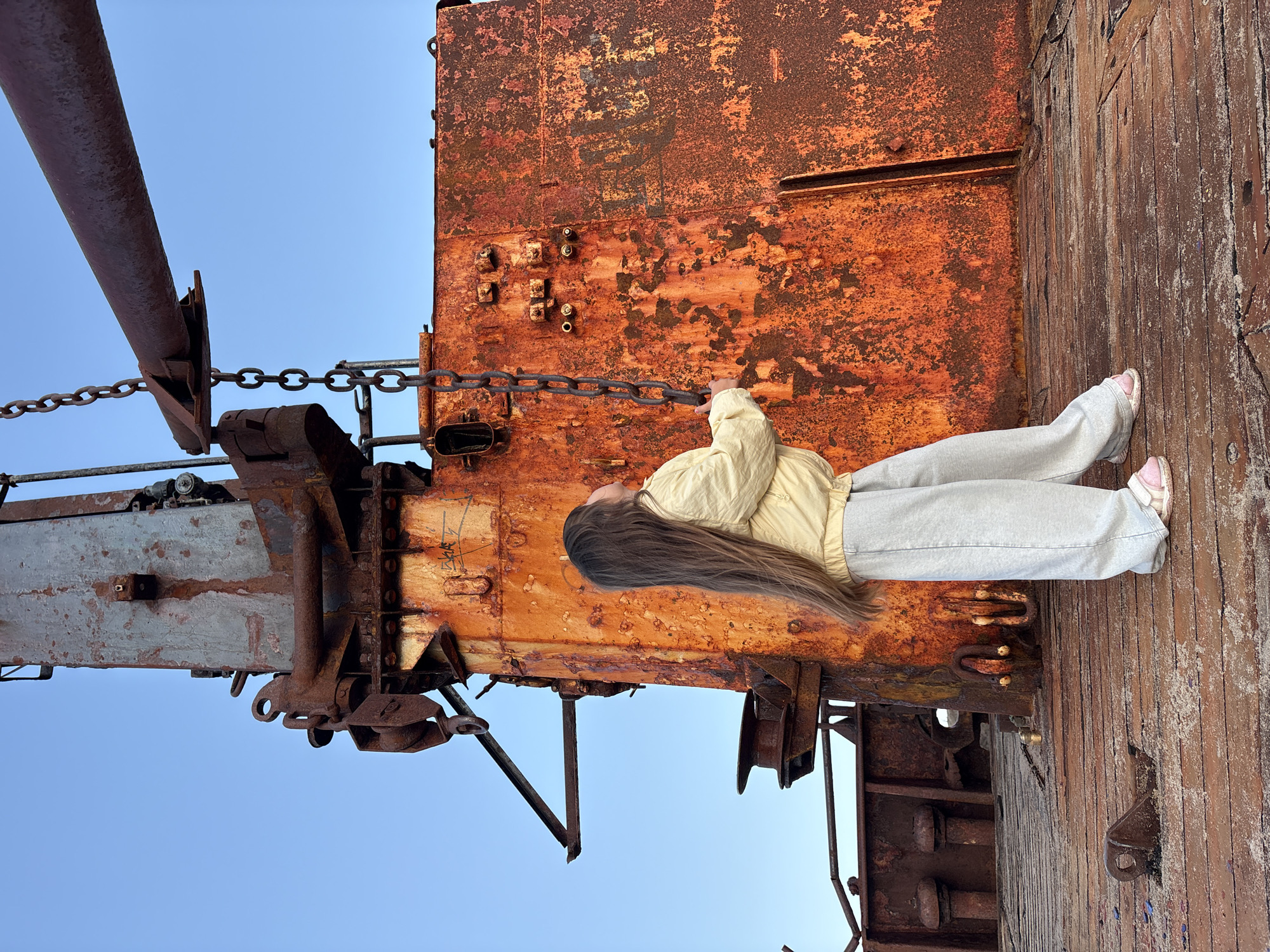 A young girl with long brown hair, wearing a light yellow jacket and grey pants, stands on the weathered wooden deck of a rusty ship. She is looking up at a thick metal chain hanging from a large, corroded industrial crane. The crane and the ship's structure are covered in a vibrant orange rust, contrasting with the clear blue sky. The scene appears to be set in the city of Teriberka, Russia, a location known for its ship graveyards and harsh coastal environment. The ground beneath her feet is also made of old wooden planks, showing signs of decay and wear.