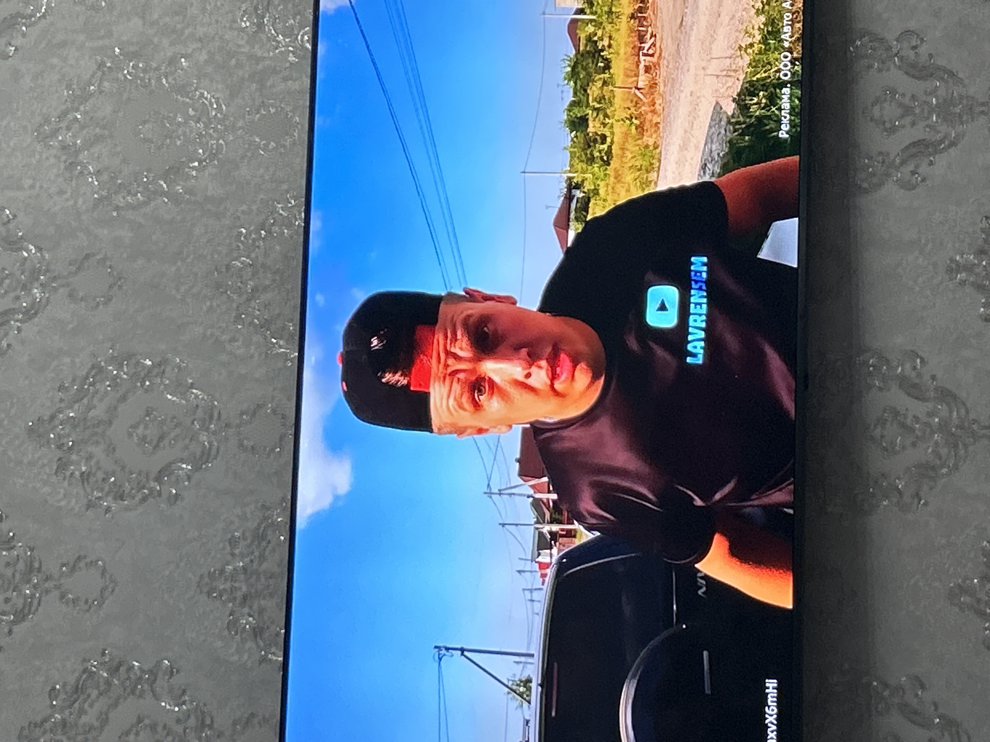 The image displays a television screen showing a man wearing a black t-shirt and a black baseball cap with a red brim. He is outdoors, with a clear blue sky and some clouds visible. In the background, there are houses, trees, and utility poles with wires. A dark-colored SUV is partially visible on the left side of the screen. A logo with a play button and the text "LAVRENSFM" is displayed on the man's t-shirt. Text in Russian, "Реклама. ООО «Авто А", is present in the bottom right corner of the screen, indicating an advertisement. The image quality suggests it is a photograph of a screen, with some glare and wallpaper visible around the screen. There is no indication of the specific location within Khalimbekaul, Russia.