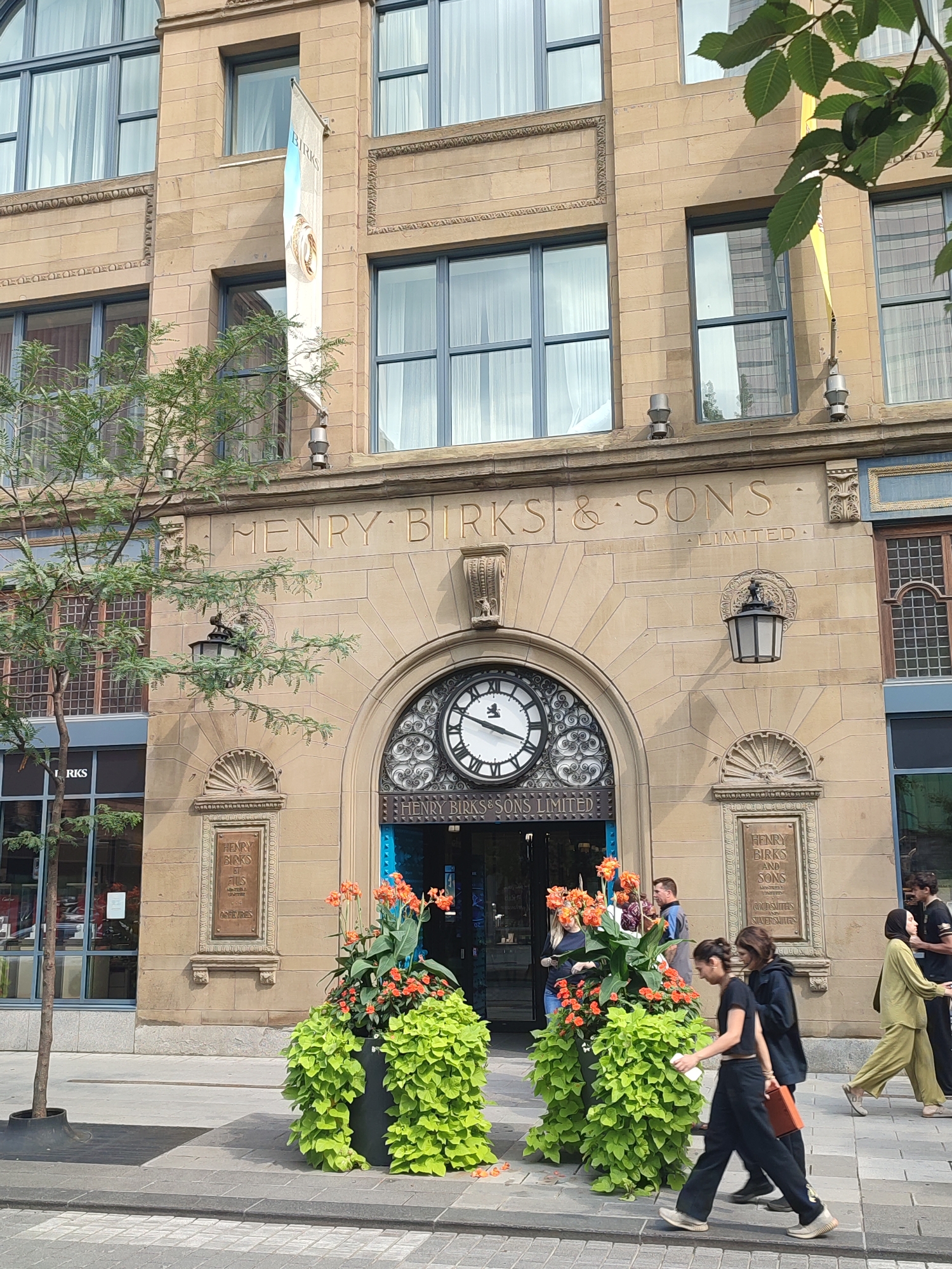 The image depicts the exterior of the Henry Birks & Sons building in Montreal, Canada. The building is constructed of light-colored stone with classical architectural elements, including ornate carvings above the entrance and windows. Above the arched entrance, the words "HENRY BIRKS & SONS LIMITED" are carved into the stone. A large, decorative clock is prominently featured within the archway.
In front of the entrance, large planters filled with bright green foliage and orange flowers add a splash of color. Several individuals are visible on the sidewalk in front of the building. Two women are walking towards the right, one carrying an orange item. Another couple, a man and a woman wearing a hijab, are walking in the same direction on the far right. A man wearing a blue shirt stands near the planters.
The building has large windows with reflective glass, some of which are framed by classical details. A white banner with the word "BIRKS" and an image of rings hangs vertically on the facade to the left of the entrance. A young tree with sparse foliage stands on the left side of the sidewalk. The sidewalk is paved with gray stones, and a crosswalk is visible in the foreground. The overall scene captures a well-maintained urban environment with a focus on the historic Birks building.