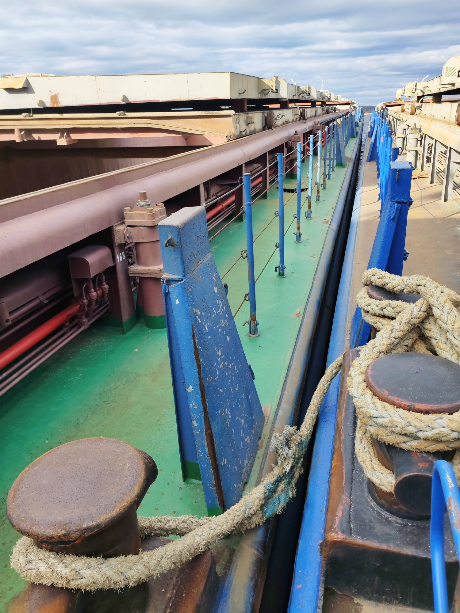 The image depicts a close-up view of a section of a large, industrial barge or ship, potentially docked. The scene is dominated by heavy metal structures, including pipes, railings, and mooring bollards with ropes. The deck is painted in a bright green color, and the railings are painted in blue. The mooring bollards are black with rust-colored tops. A thick, light-colored rope is wrapped around the bollards. The background features more of the barge's structure, including what appears to be heavy machinery and additional metal plating. The sky is overcast.
While it is impossible to confirm the location definitively as Rybinsk, Russia without further evidence, the industrial nature of the barge and the overcast sky are consistent with such a location.