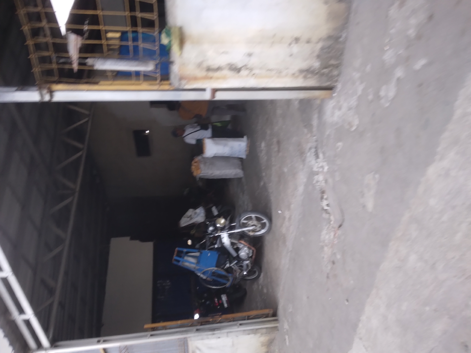 The image depicts an outdoor scene in Desa Pamijen, Kecamatan Sokaraja, Indonesia. In the foreground, a concrete ground is visible with some debris. A structure with a metal roof and support beams forms a covered area. Inside this structure, a motorcycle with a blue cage-like attachment on its back is parked. Behind the motorcycle, two large white sacks are present, possibly containing goods. Further in the background, two individuals are standing. One is wearing a white shirt and dark pants, and the other is wearing an orange shirt and dark pants. The environment appears to be utilitarian, possibly a storage area or workshop.