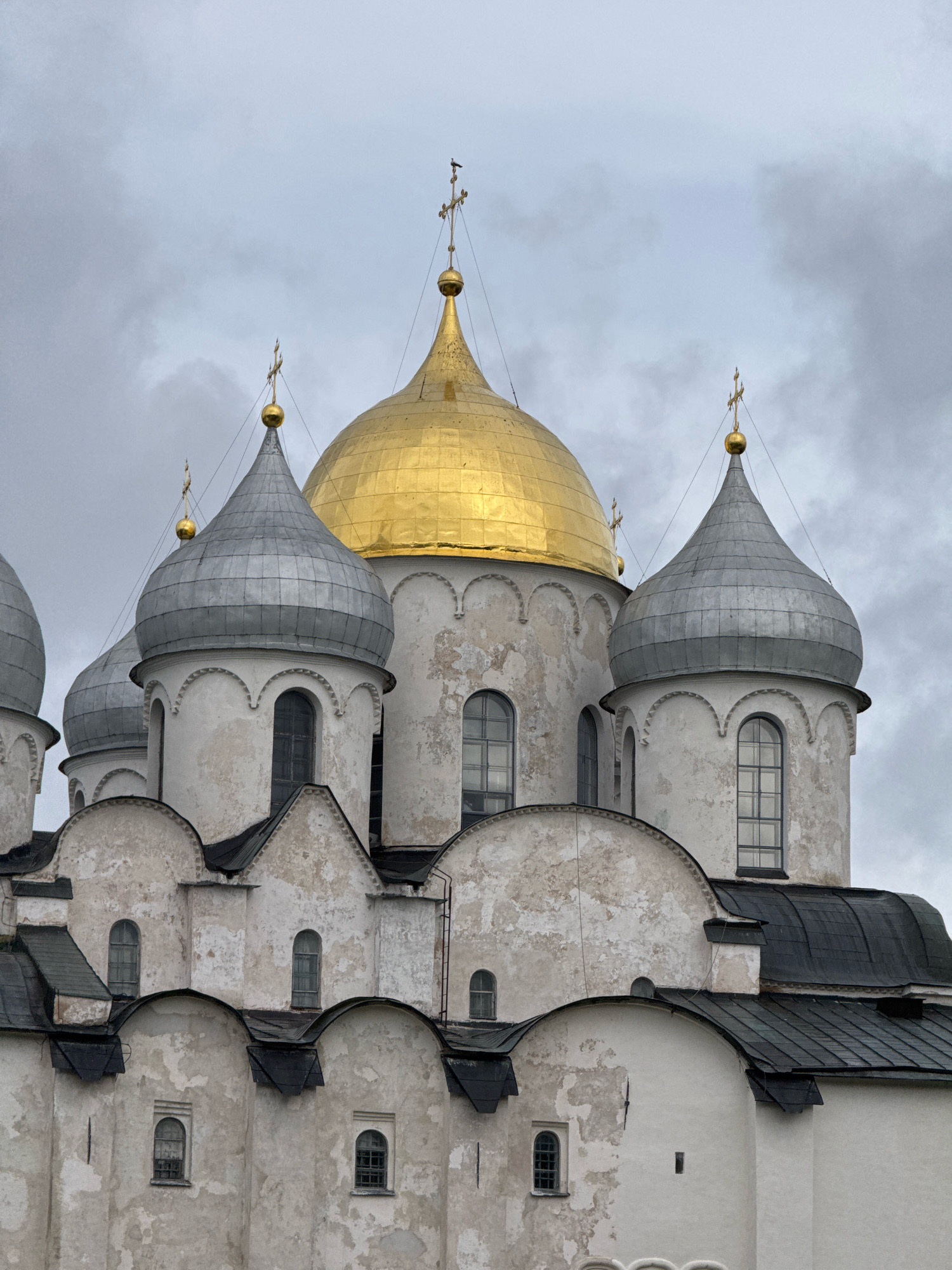 The image shows a close-up view of Saint Sophia Cathedral in Veliky Novgorod, Russia, under a cloudy sky. The cathedral features multiple domes, including a prominent golden dome at the center and several silver, onion-shaped domes. The white exterior walls of the cathedral are weathered, showing patches of plaster that have worn away, revealing the brickwork underneath. Small, arched windows are visible on the different levels of the structure. Dark, sloped roofs cap various sections of the building. The overall scene is dominated by the architecture of the cathedral, with the cloudy sky providing a muted backdrop. There are no people or visible actions occurring in the image.