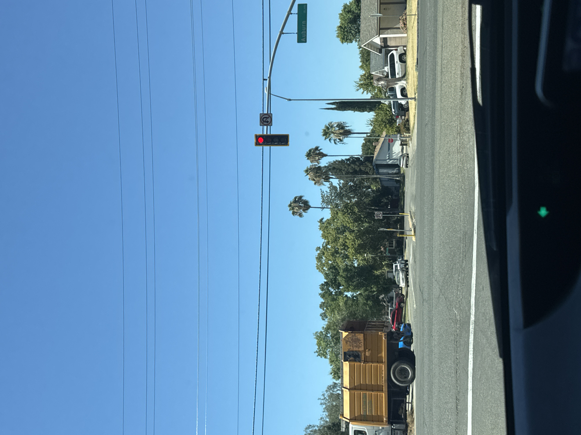 The image captures a street scene in Citrus Heights, USA, on a clear, sunny day. In the foreground, the dashboard of a vehicle is visible, suggesting the photo was taken from a car. The road surface is asphalt with clear lane markings.

A large yellow garbage truck is parked on the left side of the road, its side visible to the viewer. Further down the road, several cars are present, including a red sedan, a white SUV, and a light-colored sedan.

A traffic light is suspended over the intersection, displaying a red light. A sign indicating "Auburn BLVD" is mounted on a street sign pole. In the background, residential houses with beige and brown exteriors are visible, surrounded by lush green trees, including several palm trees. The sky is a bright, cloudless blue. Several utility wires are strung across the sky. A U-turn prohibition sign is also visible near the traffic light.