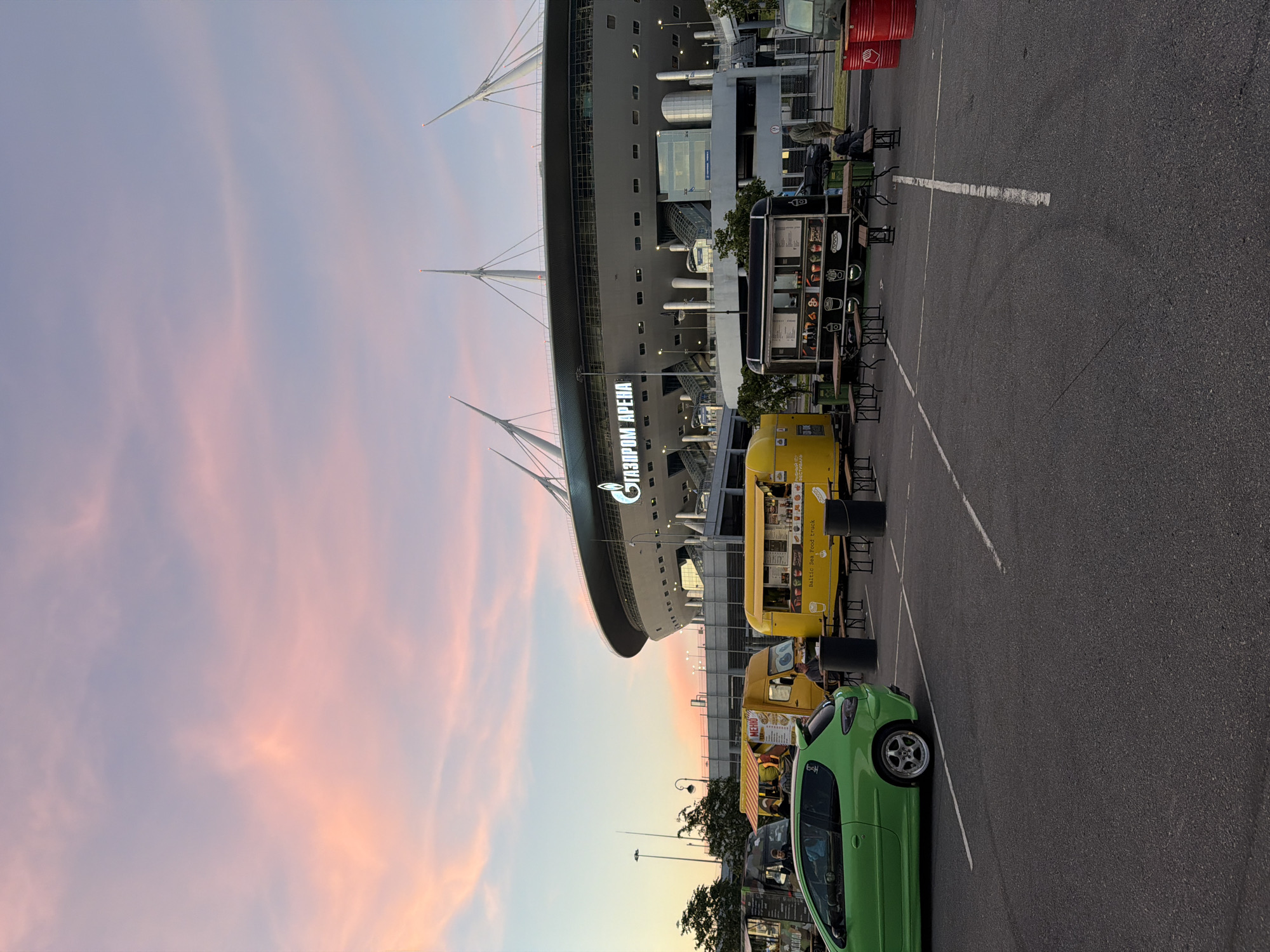 The image shows a parking lot in front of the Gazprom Arena in St. Petersburg, Russia, during sunset. Several food trucks are parked in the lot, including yellow trucks and a black truck with the words "Baltic Sea Food truck" visible on it. A green car is parked in the foreground. People are visible around the food trucks, some standing and some sitting at picnic tables. The sky is filled with soft pink and purple clouds, indicating the sun is setting. The Gazprom Arena stadium is visible in the background, with its distinctive pointed roof.