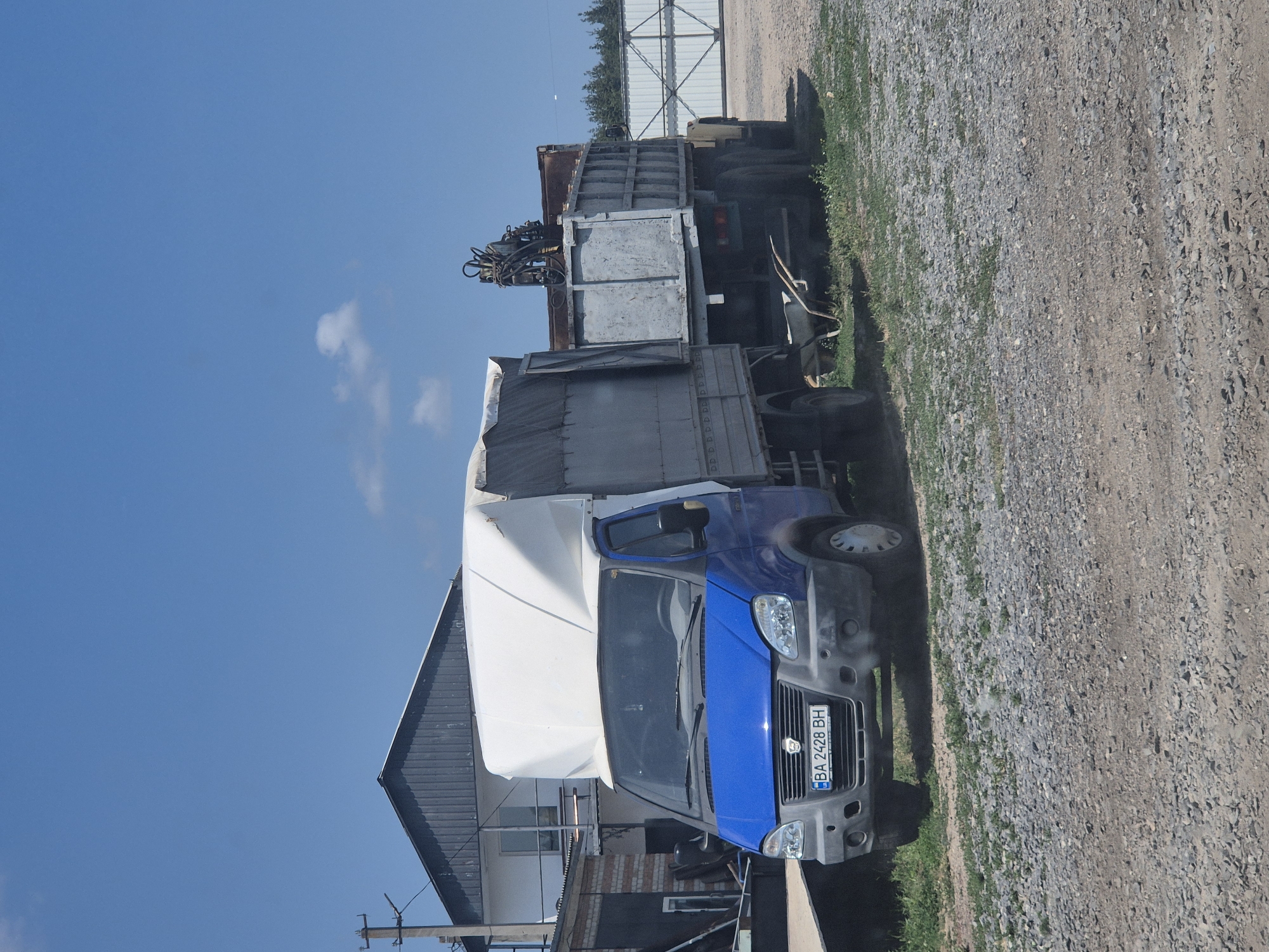 A blue truck with a white tarp-covered cargo area and a grey trailer are parked on a gravel lot in Pidhaitsi, Ukraine. The truck has a license plate that reads "BA 2428 BH". The trailer is a dump trailer with a hydraulic crane. In the background, there is a building with a dark metal roof and a white section. A white fence is visible behind the trailer. The sky is clear and blue with a few small clouds. The scene appears to be a sunny day in an industrial or rural area. There are no people visible in the image.