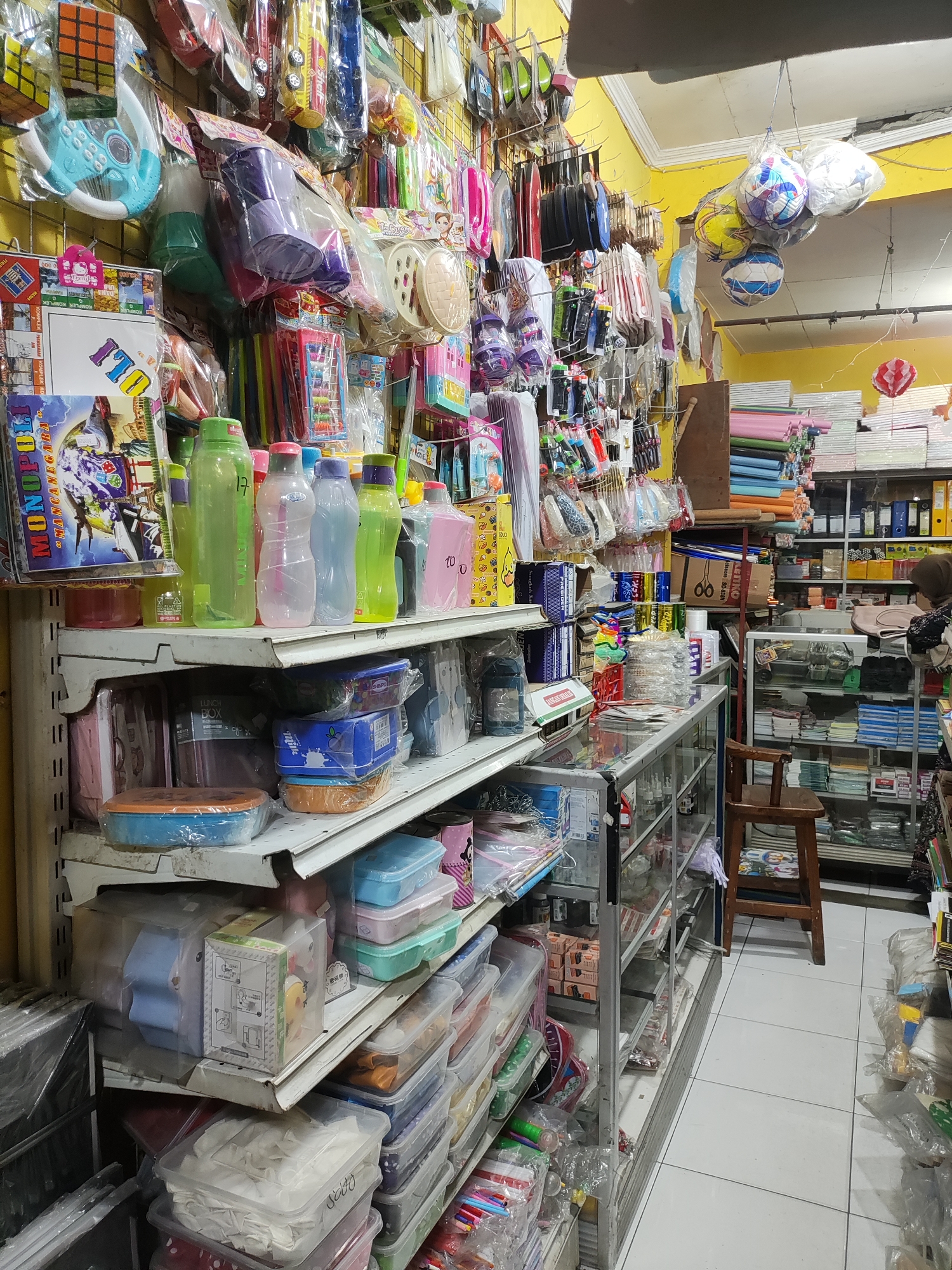 The image depicts the interior of a retail store, likely a stationery or variety store, located in Wanajaya, Indonesia. The store is densely packed with merchandise displayed on shelves and hanging from racks.
On the left side of the image, shelves are filled with various items including plastic containers, water bottles of different colors, and what appear to be educational toys or games such as a Rubik's cube and a toy steering wheel. Boxes of Monopoly are visible on a higher shelf.
The middle section of the store features more shelves stocked with an assortment of goods, including more plastic containers, colorful stationery items like pens and pencils, and packaged toys. Several hanging displays showcase smaller items, possibly office supplies or craft materials.
The right side of the image reveals more shelving units, including a glass-fronted display cabinet containing what look like office supplies and possibly some snacks or small retail items. A woman with a headscarf is partially visible in the background, standing near a counter. Rolls of colored paper are stacked on shelves in the background. A wooden stool is placed in front of a glass display case.
The overall environment is well-lit with fluorescent lighting, and the walls are painted a bright yellow. The flooring appears to be white tiles. The store seems to be organized, but the sheer volume of products creates a crowded visual.