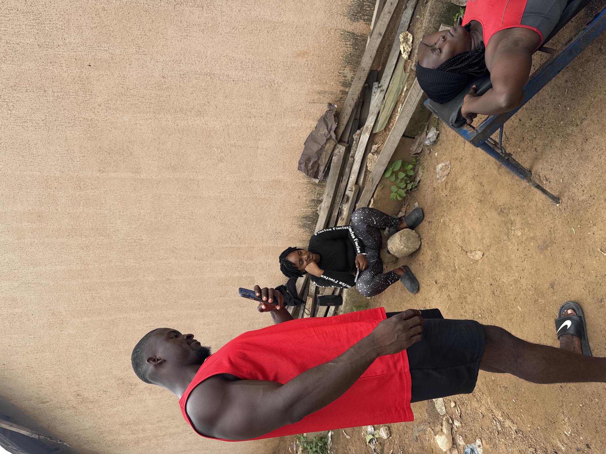 In Akwanga, Nigeria, an outdoor scene unfolds with three individuals. A man, clad in a red tank top and black shorts, stands to the left, holding a smartphone. To his right, a woman in a black long-sleeved shirt and patterned leggings sits on a log, looking down at her hands. Further to the right, another woman lies on a blue workout bench, eyes closed, wearing a red and gray tank top and a black head covering. The environment is a dirt-covered area with a textured, pale wall as a backdrop. Scattered debris, including plastic bags and pieces of wood, are visible on the ground. A stack of weathered wooden planks leans against the wall behind the seated woman.