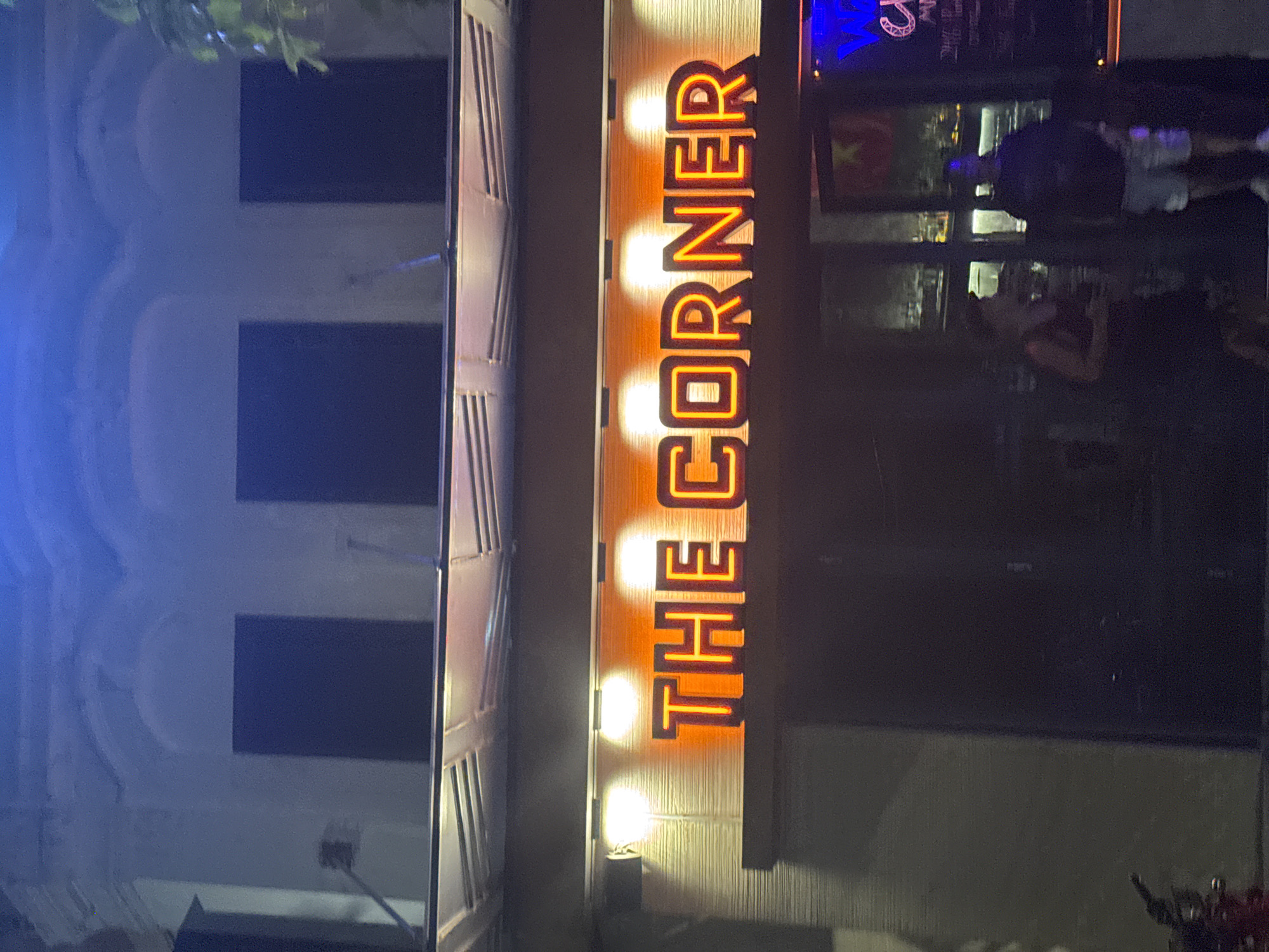 A vertical neon sign reads "THE CORNER" in glowing orange letters against a wooden background. Three spotlights illuminate the sign from below. To the left of the sign, the exterior of a building is visible, featuring dark rectangular windows and architectural details that suggest an older building. The windows reflect a blue light, possibly from streetlights or interior lighting. To the right of the sign, the scene transitions to a darker area with reflections in a glass surface. These reflections show glimpses of people and the interior of a building, including what appears to be a Vietnamese flag. The overall lighting suggests it is nighttime. The location is identified as Hà Nội, Vietnam.