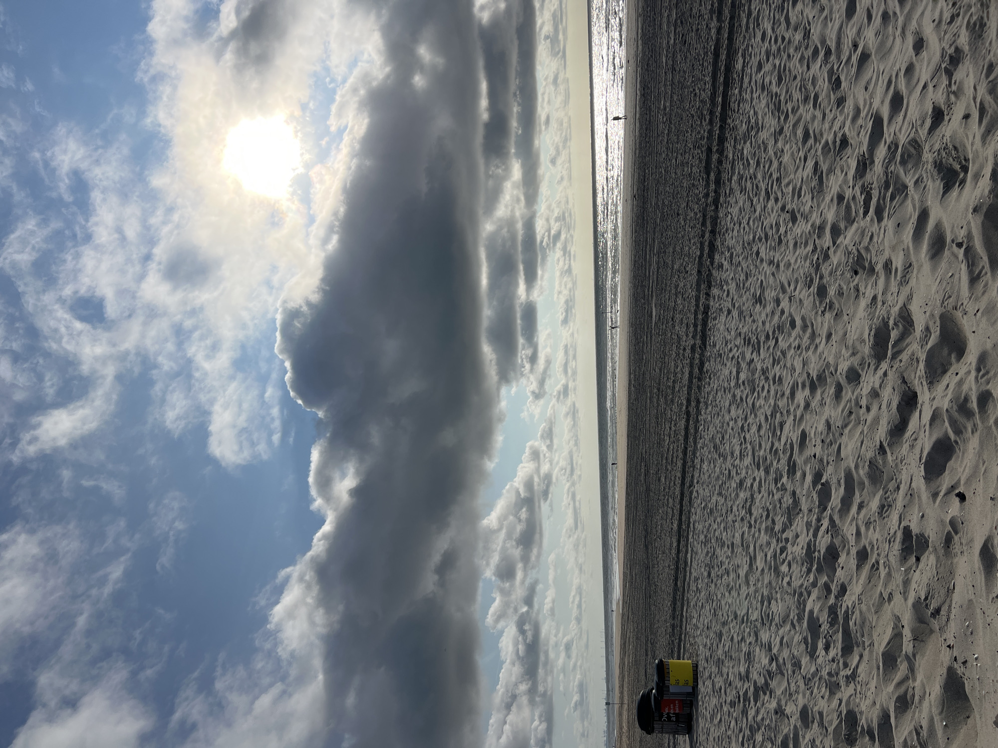 This is a wide-angle, eye-level, vertical photograph of a beach in The Hague, Netherlands. The foreground is dominated by sand dunes and the texture of the sand. In the mid-ground, the beach transitions to wet sand and then the ocean. The ocean is calm with small waves and reflects the sunlight. The horizon is visible with a few distant figures walking on the beach. The sky is partly cloudy with dramatic dark and light clouds, and the sun is partially obscured by clouds, creating a bright halo effect. In the far left of the frame, two trash cans are visible. The scene is one of a typical day at the beach.
