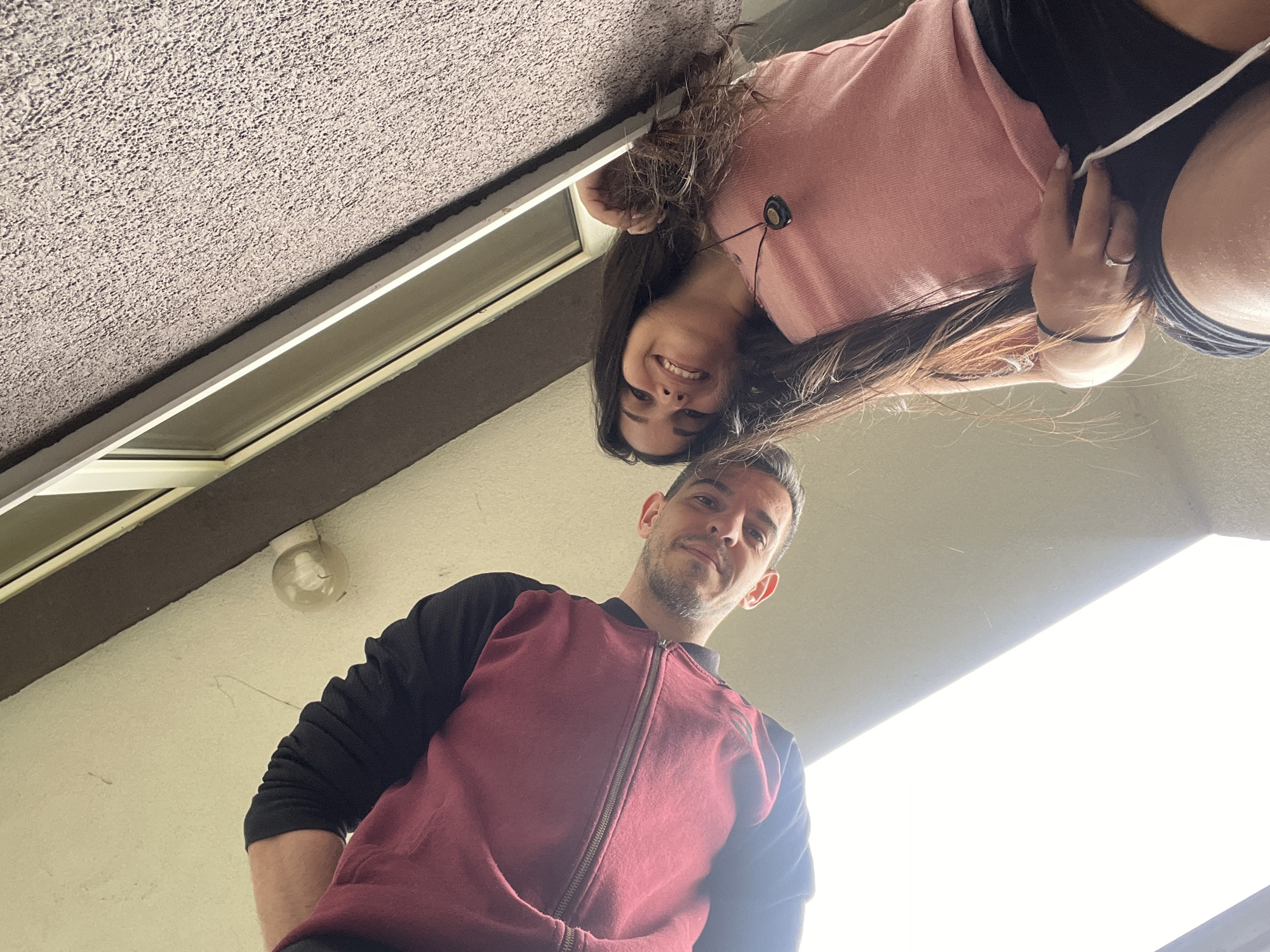 Two individuals, a male and a female, are captured in a low-angle, upward-facing selfie. The male, on the left, wears a maroon zip-up jacket with black sleeves. He has short, dark hair and a stubble beard, and is looking down at the camera with a slight smile. The female, on the right, has long, dark hair and wears a pink tank top and black shorts. She is smiling and looking towards the camera, her right hand resting on her thigh. A dark pendant necklace is visible around her neck. They are positioned outdoors, with a textured, light-colored wall and a section of a window visible in the background, suggesting an urban setting. The image was taken in the city of Sofia, Bulgaria.