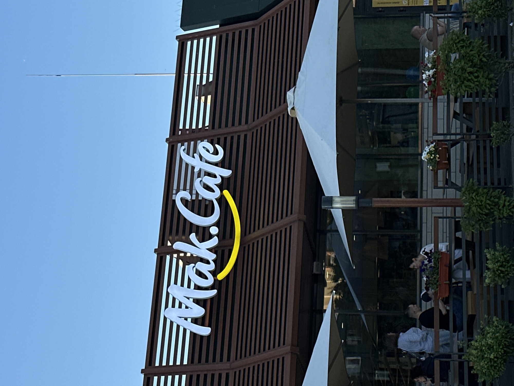 An exterior shot shows the "Mak.Cafe" sign in white lettering with a yellow smile beneath it. The sign is mounted on a brown wooden slatted facade of a building. Below the facade, outdoor seating is visible, with tables and benches under large white umbrellas. Several people are seated at the tables, some interacting with each other, while others appear to be alone. Some are obscured by the umbrellas. Green bushes are planted in front of the seating area. The sky is a clear blue. The scene appears to be in a cafe or restaurant setting.
