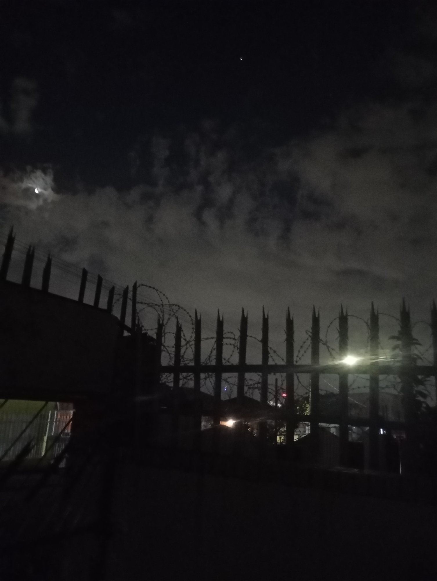 The image depicts a nighttime scene in what appears to be an urban or suburban environment within Johannesburg, South Africa. The foreground is dominated by a dark, formidable security barrier. This barrier consists of a solid wall or gate structure on the left, transitioning into a metal fence with numerous sharp, upward-pointing spikes on the right. Multiple coils of razor wire, consistent with concertina wire, are affixed along the top of the spiked fence.
Beyond the barrier, the silhouettes of buildings or houses are dimly visible against a dark backdrop. A bright, circular light source, likely a streetlamp, is prominent in the mid-right section, casting a significant glare above the fence line. Other faint artificial lights, including a small red beacon, are discernible further in the background. Sparse, dark vegetation is also visible behind the fence on the right.
The upper half of the frame shows a dark night sky with patchy cloud cover. A distinct, slender crescent moon is visible in the upper-left quadrant through a break in the clouds. A single, small, bright point of light, consistent with a star or planet, is present in the upper-middle sky section. No individuals or discernible activities are present in the scene.