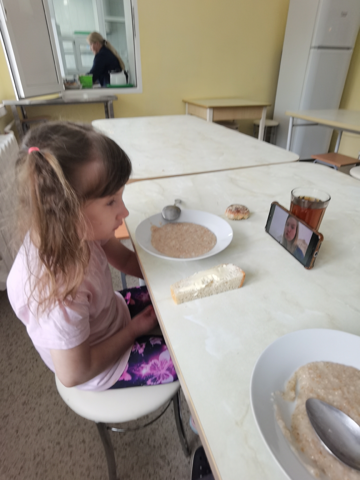 A young girl with light brown hair in pigtails, wearing a pink shirt and floral leggings, sits on a white stool at a light-colored table. On the table, there are two white plates of porridge with spoons, a slice of buttered bread, a pastry, a glass of tea, and a smartphone displaying a video call of a woman. Through a window, another woman can be seen in the background. A refrigerator is visible in the background. The location is possibly a cafeteria, the city of Perm, Russia.