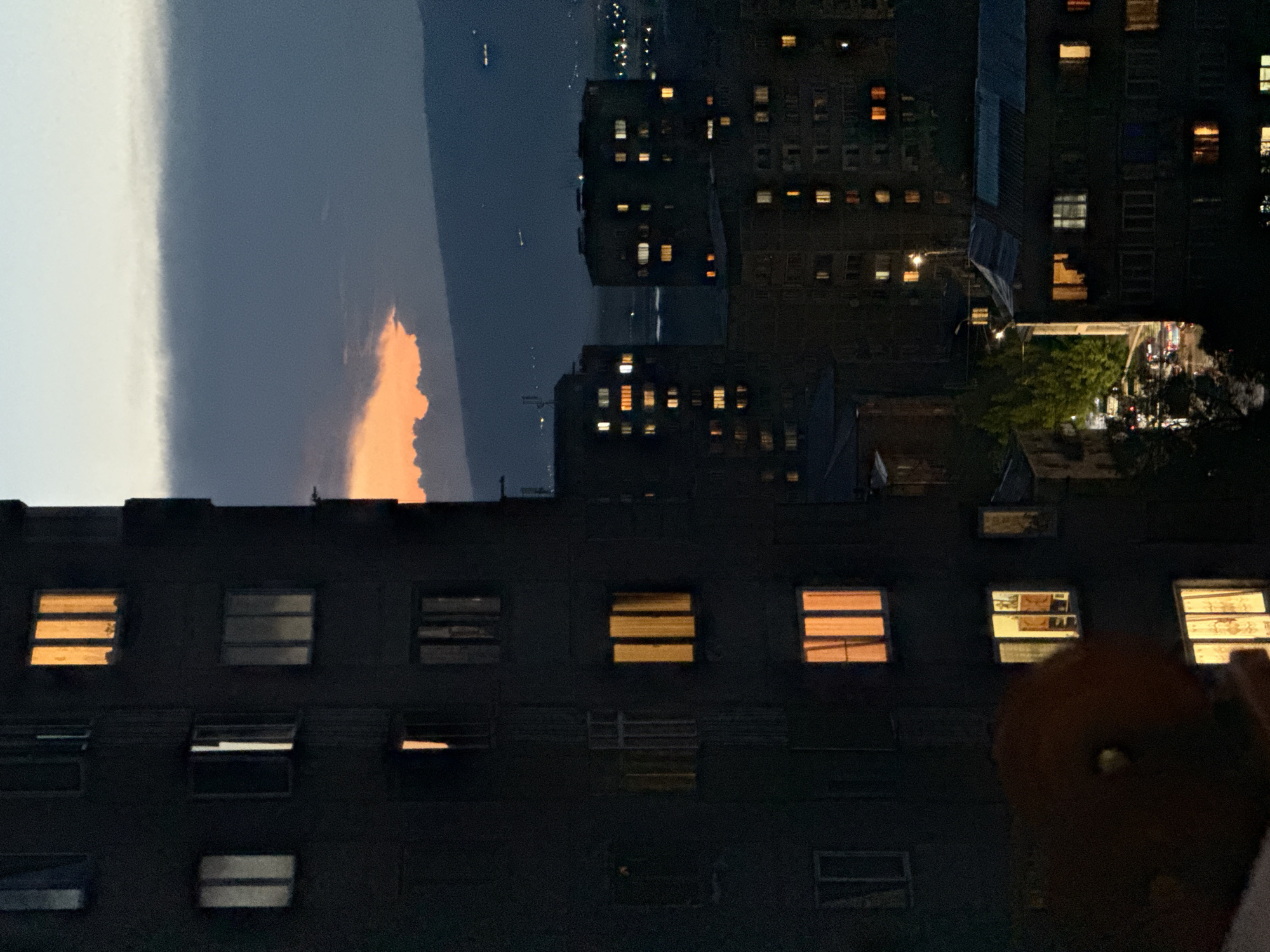The image captures a city skyline at dusk, likely in Abovyan, Armenia, with a prominent apartment building in the foreground on the left. The building's dark facade is punctuated by numerous windows, some illuminated by warm, golden light, suggesting occupancy. A few windows are dark or dimly lit. The sky above is a gradient of fading daylight, with a band of dark clouds obscuring a portion of a vibrant orange sunset. In the background, several other apartment buildings are visible, also displaying a scattering of lit windows, indicating a residential urban environment. A street with car lights is visible in the middle ground, and distant city lights twinkle on the horizon, suggesting a larger urban sprawl beyond the immediate foreground. The overall atmosphere is one of a quiet evening settling over a densely populated area.