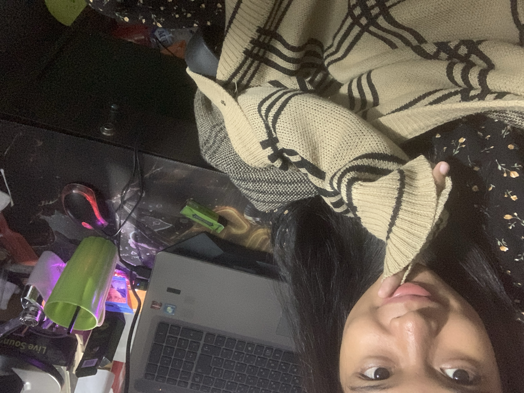 A person with long, dark hair is close to a laptop. The person is wearing a tan cardigan with black stripes over a dark floral dress. In the foreground is a black desk with various objects, including a green cup, a red computer mouse, and other small items. The location is indicated as Sungai Meriam, Indonesia.