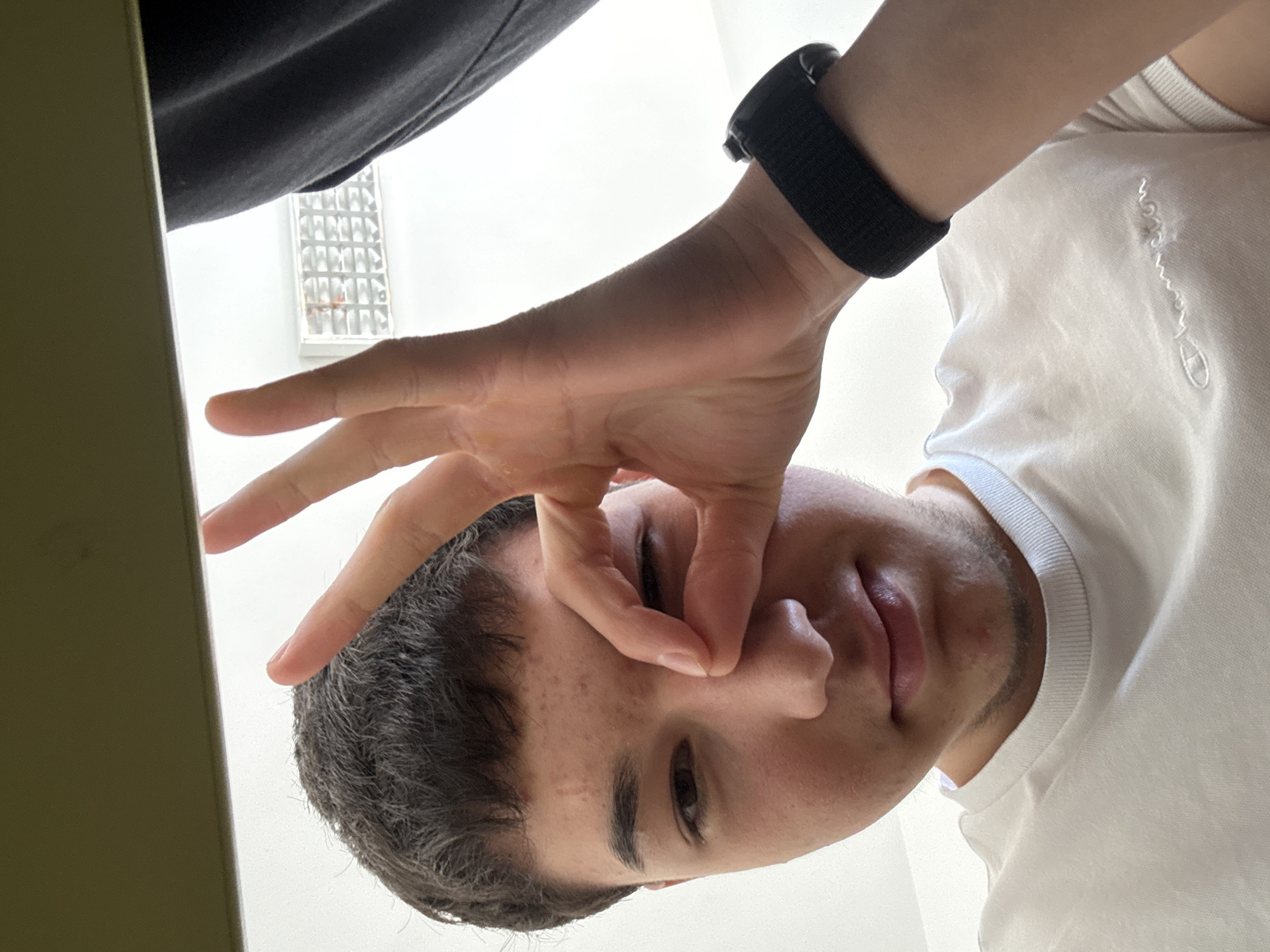 A close-up, low-angle shot depicts a person making an "OK" gesture over their right eye. The individual has short, dark hair, a light complexion, and is wearing a white t-shirt and a black smartwatch on their right wrist. The hand making the gesture is light-skinned. A portion of another person's dark-colored clothing is visible in the upper left corner. In the background, a wall with a patterned window can be seen. The location is not discernible from the image.
