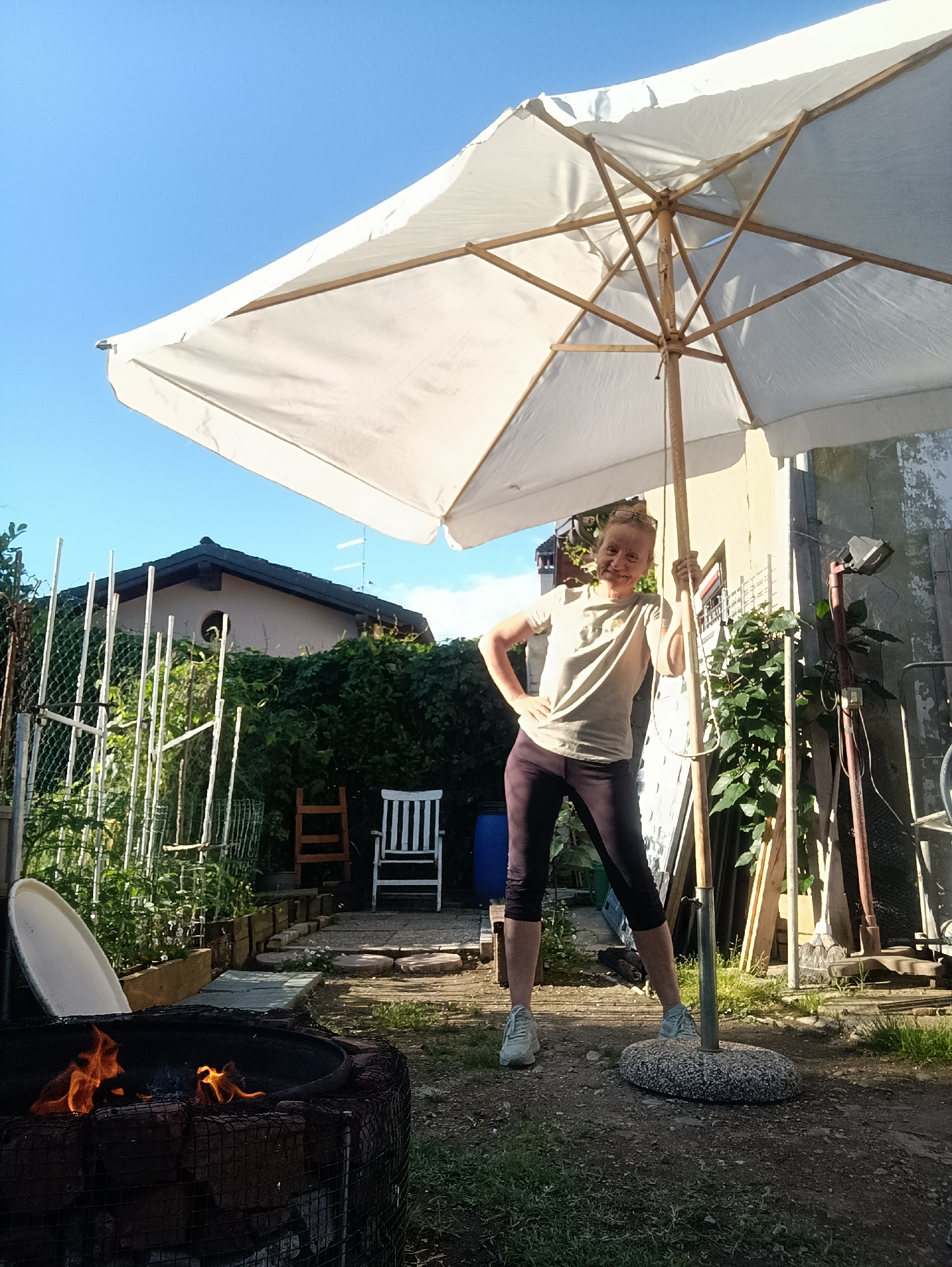 The image shows a woman standing next to a white patio umbrella in a backyard setting in Viadana, Italy. The woman is wearing a light-colored t-shirt, dark cropped leggings, and light-colored athletic shoes. She has one hand on her hip and the other resting on the umbrella pole. The umbrella is open and casting a shadow.
In the background, there's a garden area with a white fence, various plants, a white chair, and a blue barrel. A brick structure with a fire burning inside is visible in the foreground to the left. The sky is blue and clear. The surrounding environment appears to be a residential yard.