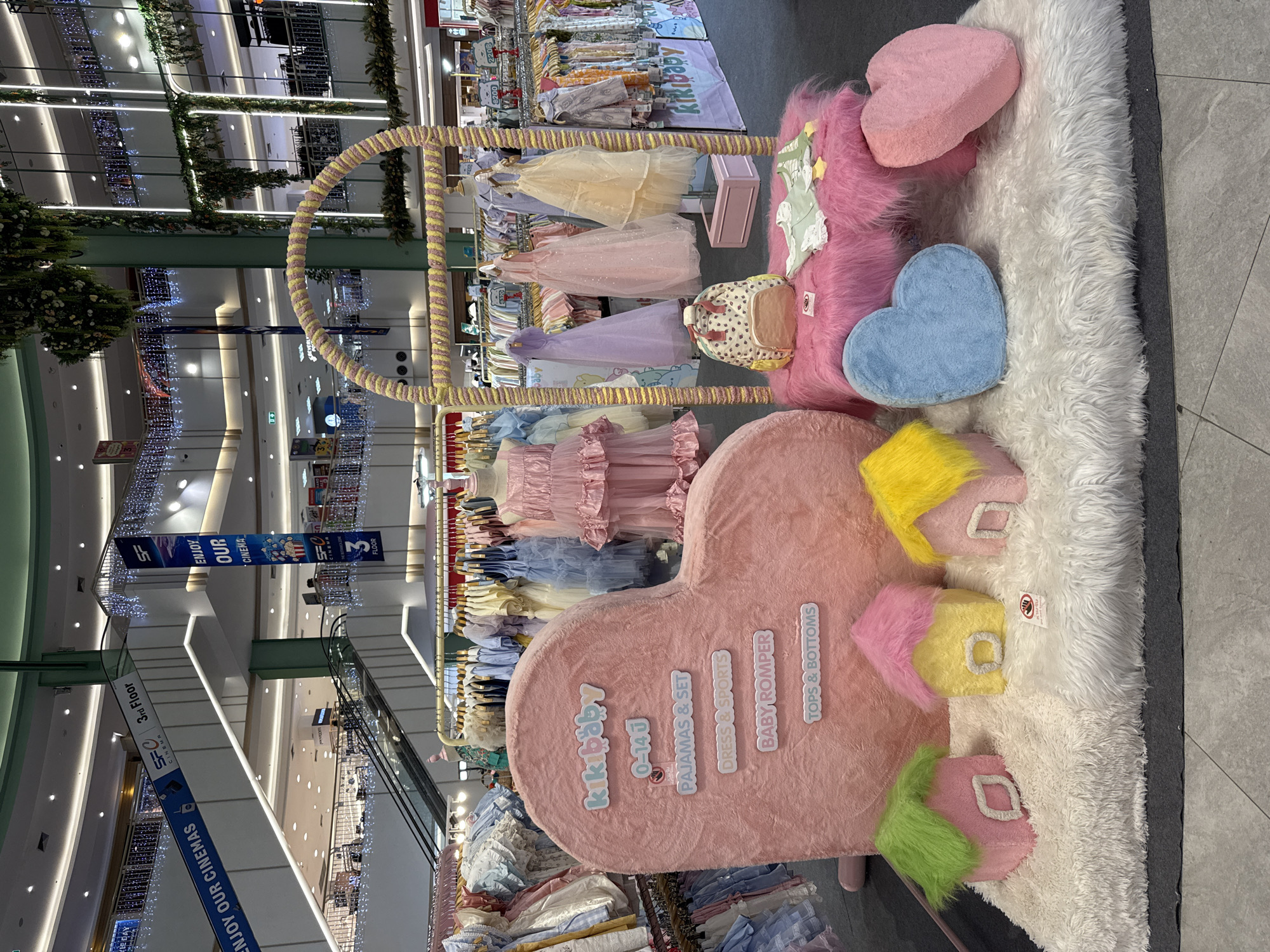 This image shows a retail display for "kikibaby" clothing inside a shopping mall in Patong, Thailand. The display includes a clothing rack with children's dresses, a pink heart-shaped sign advertising "kikibaby" products, fluffy pink and blue heart-shaped pillows, and house-shaped pillows in various colors on a white fur rug. Banners for "SFC Cinema" are visible in the background, along with escalators and other retail stores within the mall. The mall's interior includes decorative lighting and plants.